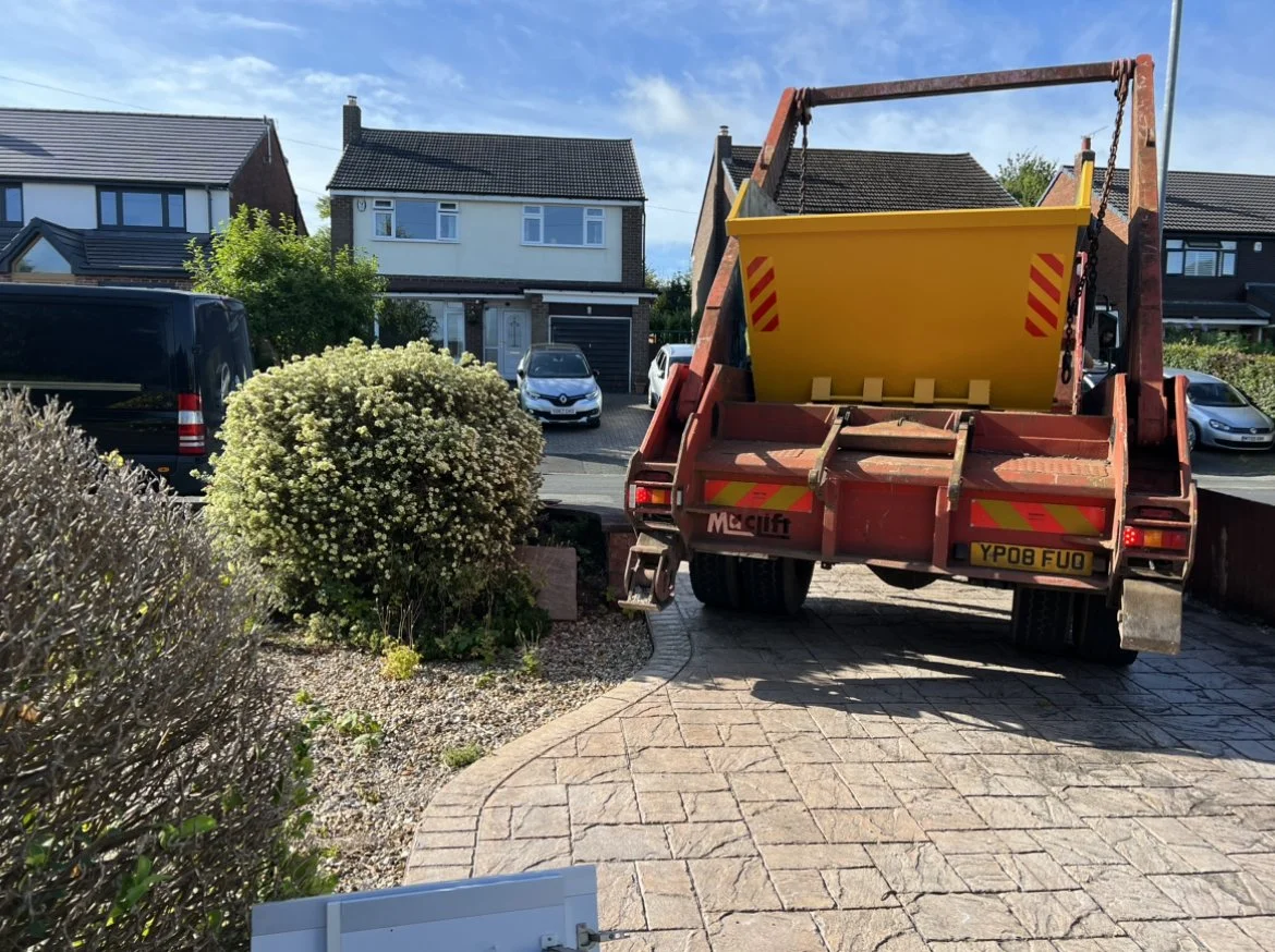 A yard with a small landscaped area containing bushes and gravel, a large yellow waste container on a red flatbed truck, and a residential street with parked cars and houses under a partly cloudy sky.
