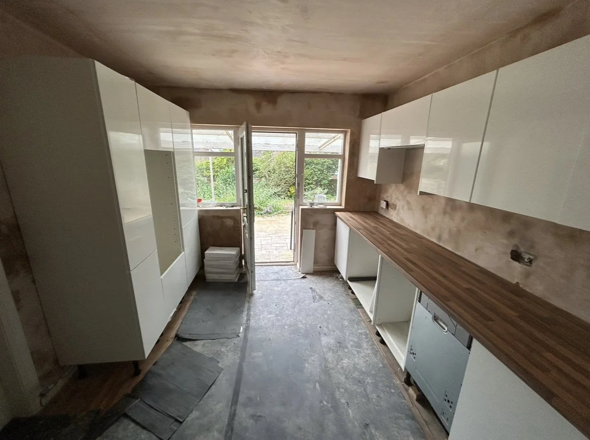 Kitchen under renovation with white cabinets, wooden countertop, window and open door leading outside.