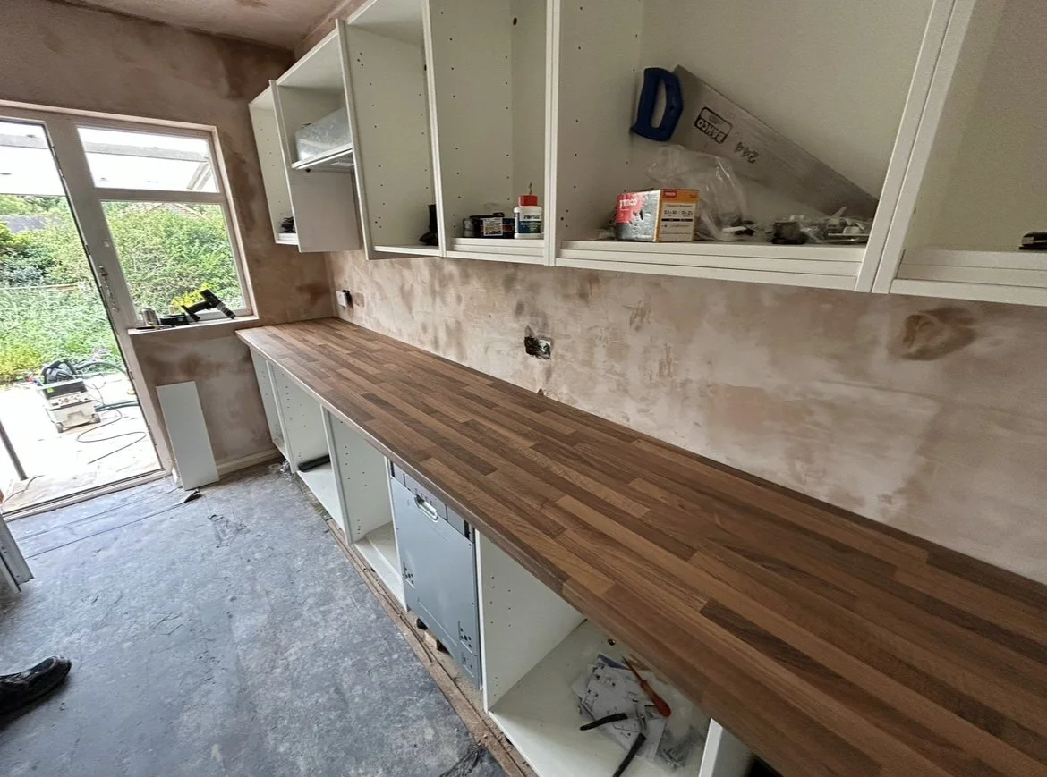 Kitchen under renovation with a long wooden countertop, unfinished white cabinets, and partially painted wall. A window shows greenery outside.