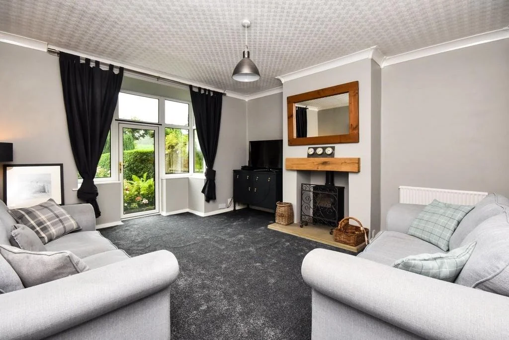 Living room with light grey walls, two grey sofas with pillows, black curtains over a large window, a small black TV stand, a fireplace with a wooden mantel, a mirror above the fireplace, decorative items, and a dark carpeted floor.
