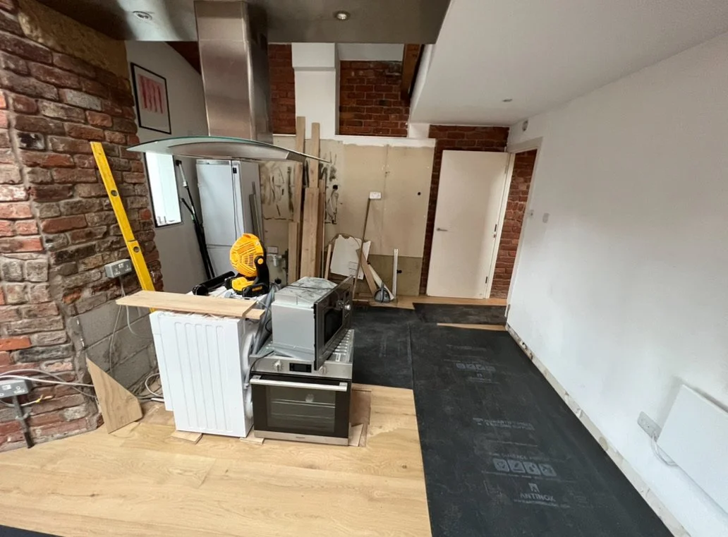 Kitchen under renovation with construction tools, wooden planks, and appliance packaging in a room with brick and white walls, a black floor covering, and a neighboring door.