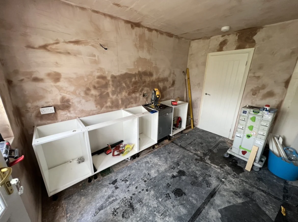 Room under construction with partially installed white shelving units, a yellow spirit level, a power saw, and tools scattered around. Unfinished walls and floors with visible dirt and marks.