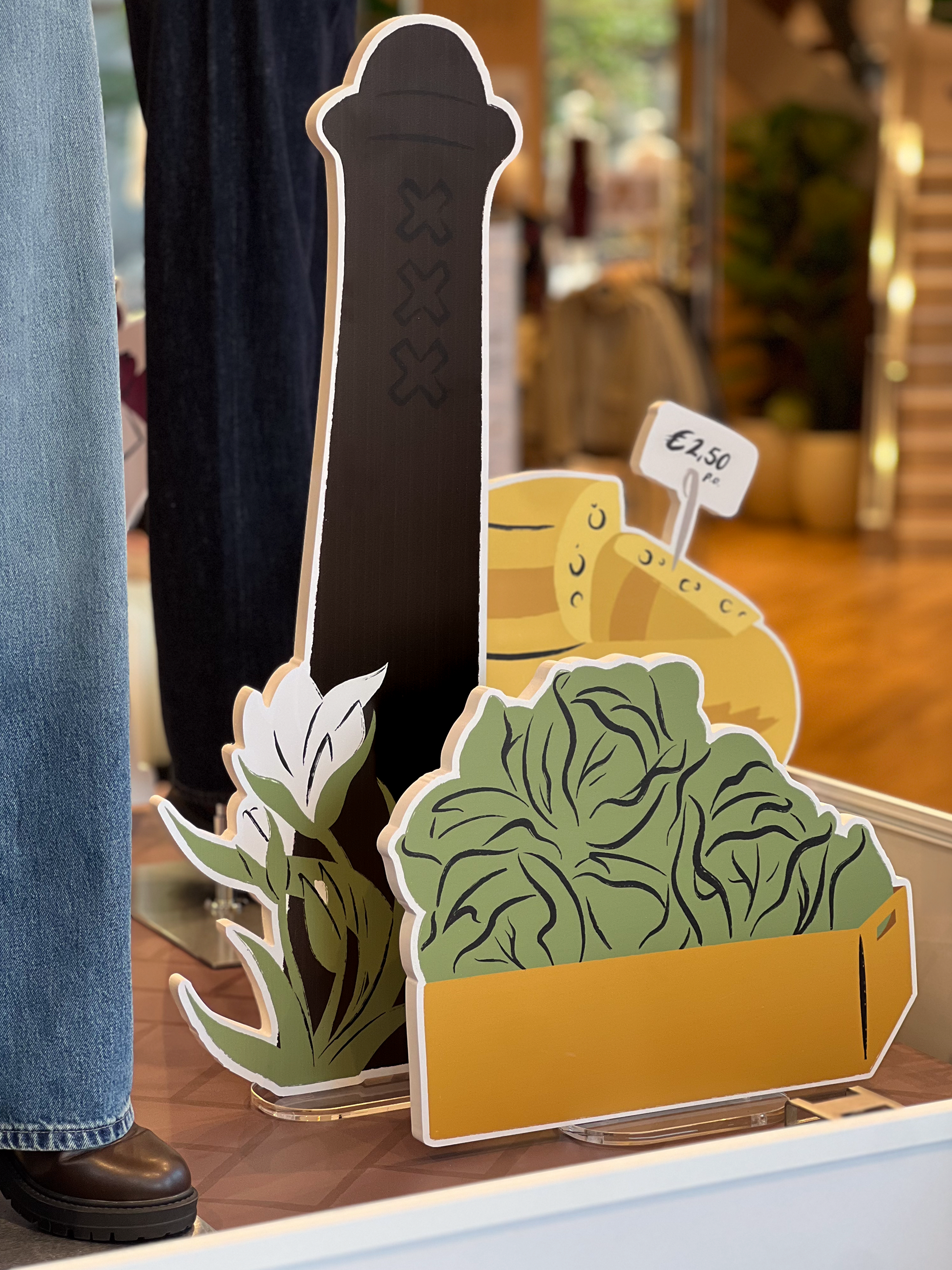 Illustrated store display by Sandy van Helden featuring stylized props of Amsterdam’s iconic bike Amsterdammertje, tulips, leafy greens in a crate, and cheese wheels, part of UNIQLO Koningsplein’s autumn 2025 market-themed visual merchandising.