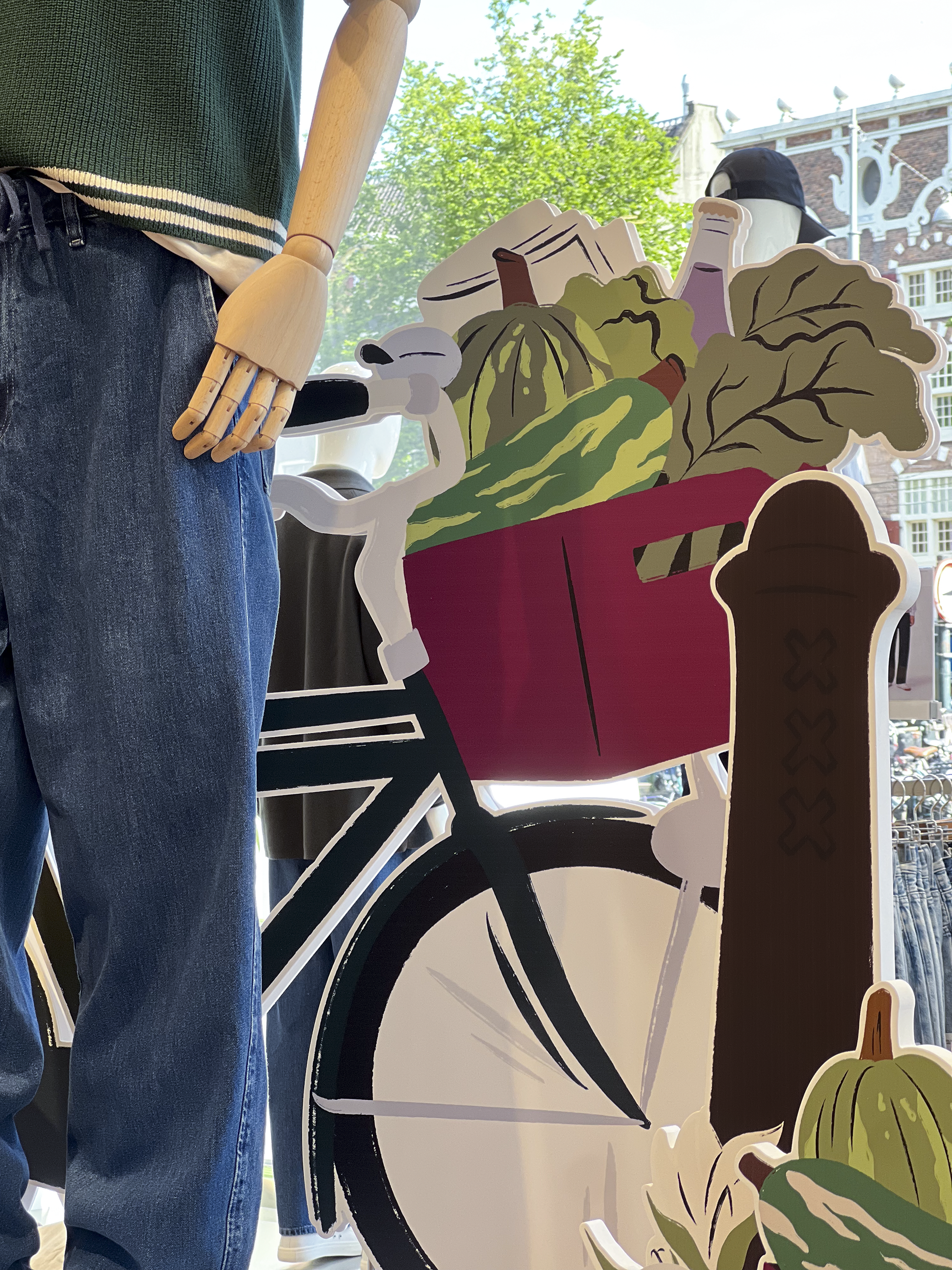 Illustrated store prop of a bike with a purple crate filled with autumn market goods—lettuce, squash, a bottle of wine, and newspapers Het Parool, illustrated by Sandy van Helden, part of UNIQLO Koningsplein’s autumn 2025 display.