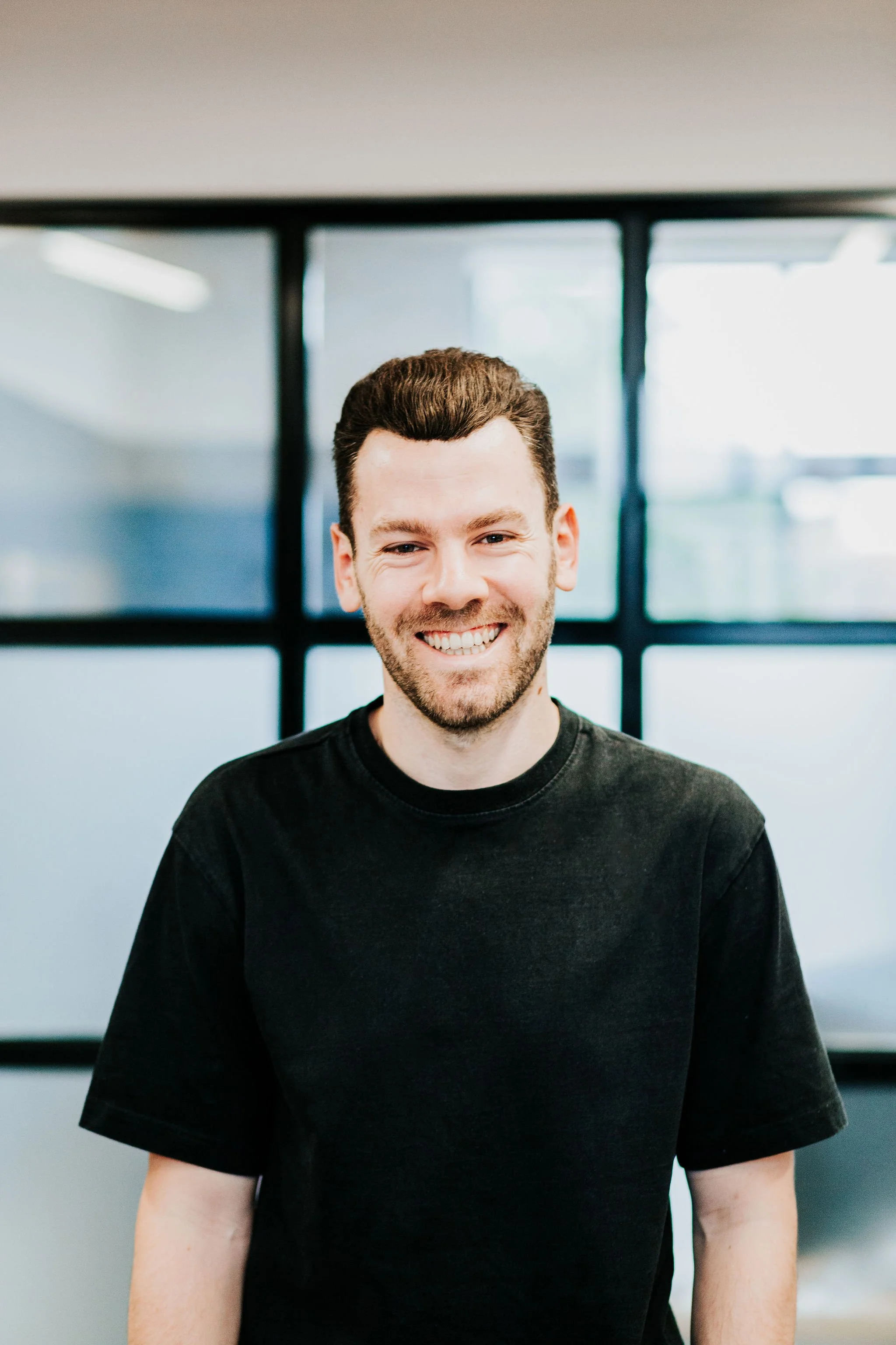 Smiling man with dark hair and beard wearing a black T-shirt, standing indoors in front of a large window with black framing.