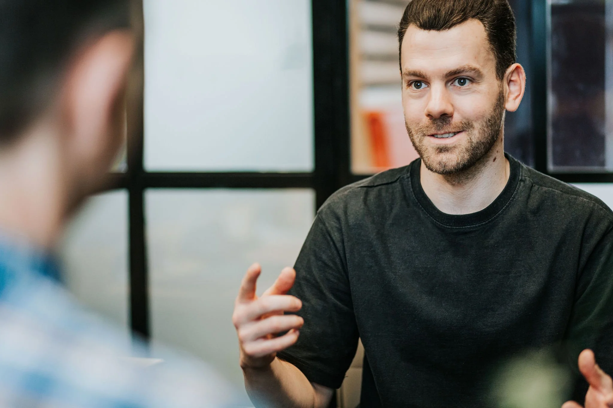 A man with dark hair and a beard, wearing a black T-shirt, is talking and gesturing with his hand while engaging in a conversation with another person in a professional setting.