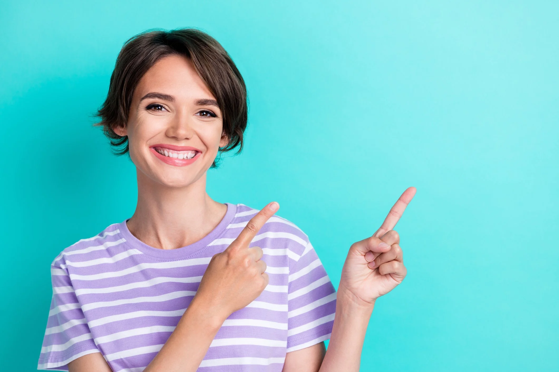 Smiling young woman with short brown hair pointing to her right, wearing a purple and white striped t-shirt, against a teal background.