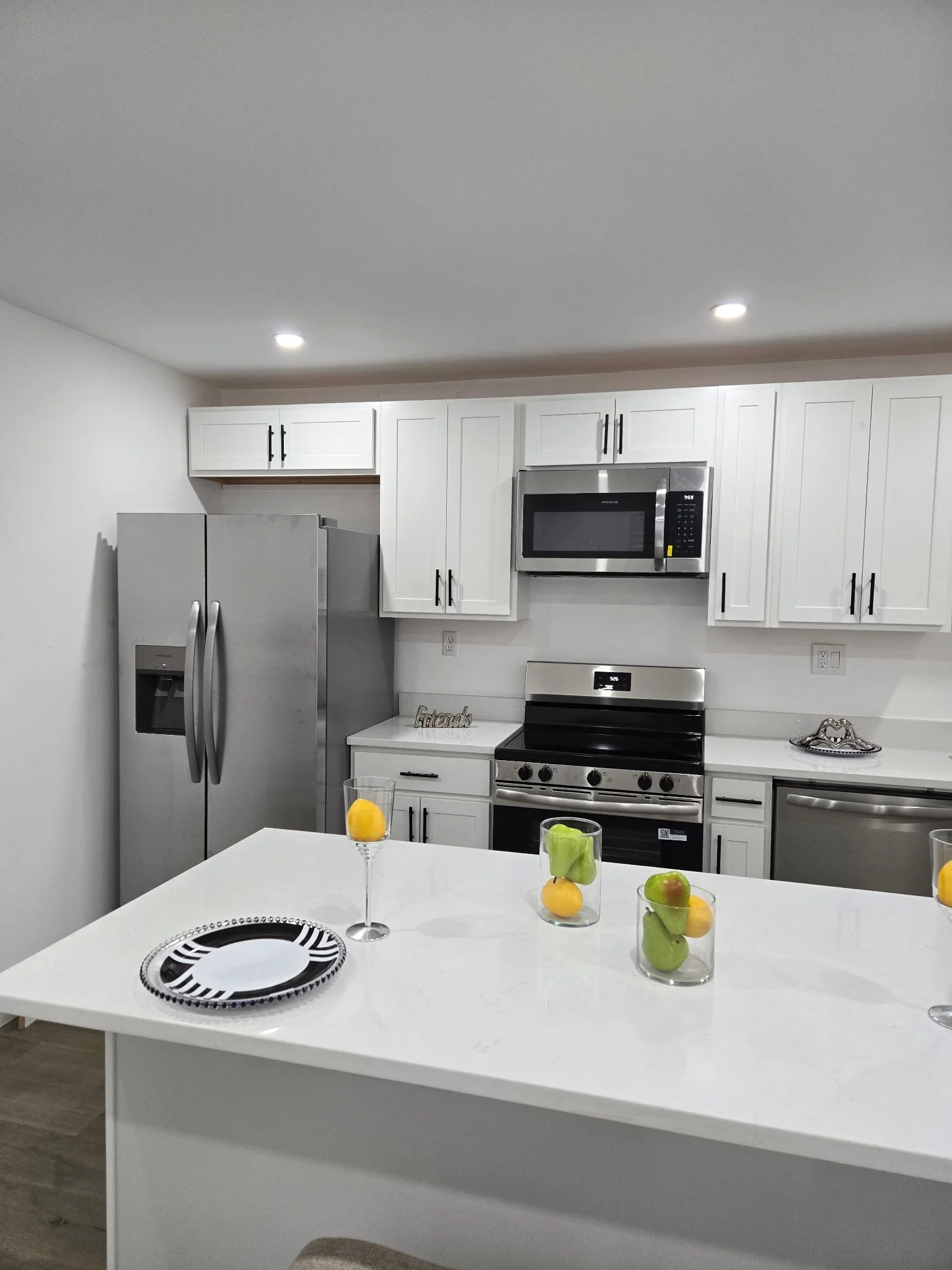 Modern kitchen with white cabinets, stainless steel appliances including a refrigerator, microwave, oven, and dishwasher, and a white island countertop with glass cups filled with fruit and a black-and-white striped plate.