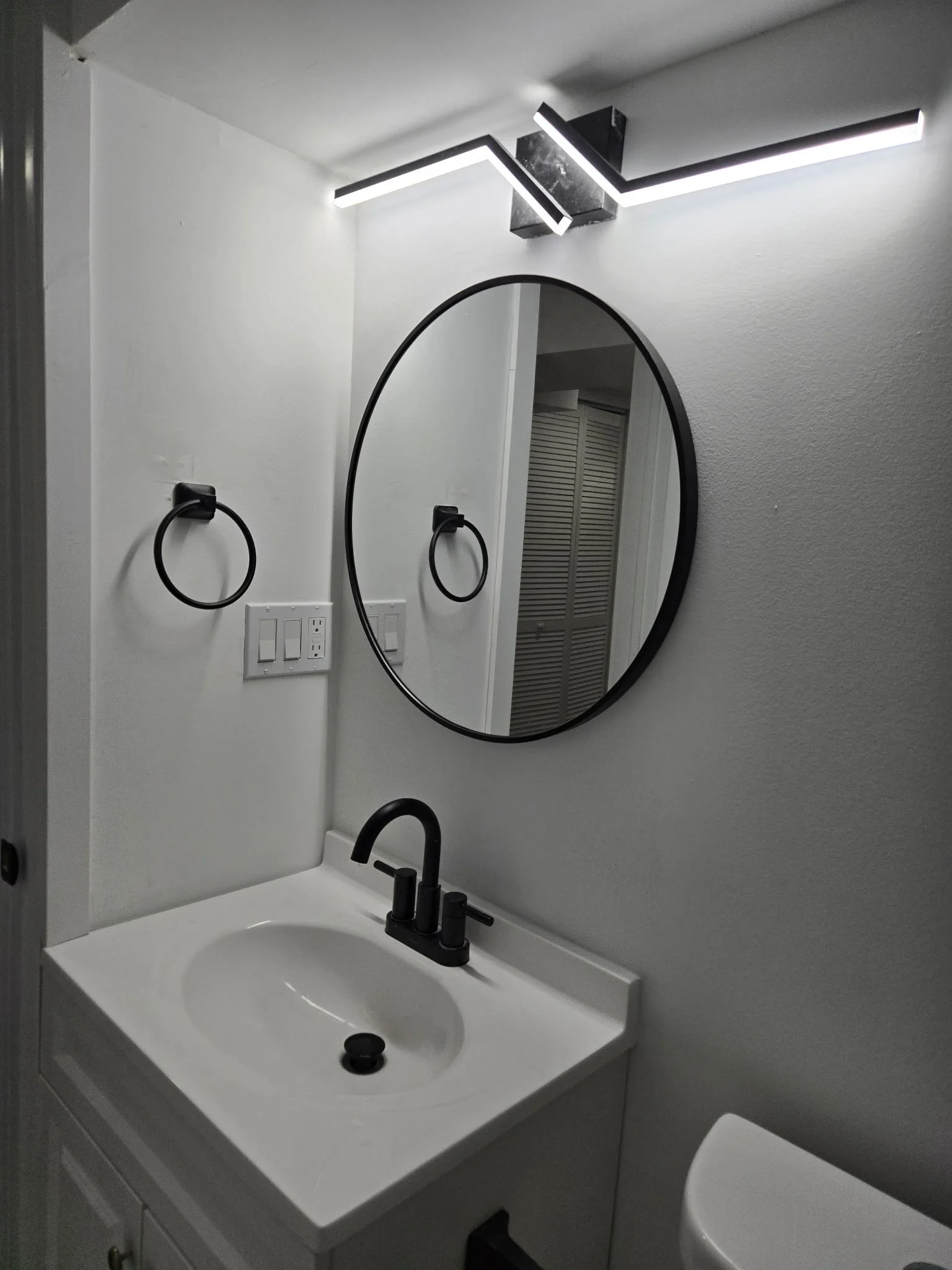 Modern bathroom with a white sink, black faucet, round mirror, black towel ring, and LED light fixture above the mirror.