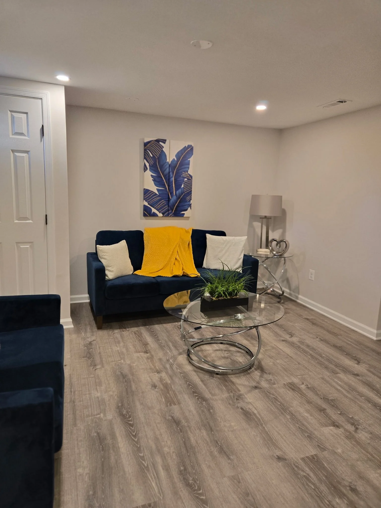 Living room with white walls, a blue velvet sofa with white and yellow throw pillows, a glass coffee table with a plant, and a side table with a lamp and decorative object.