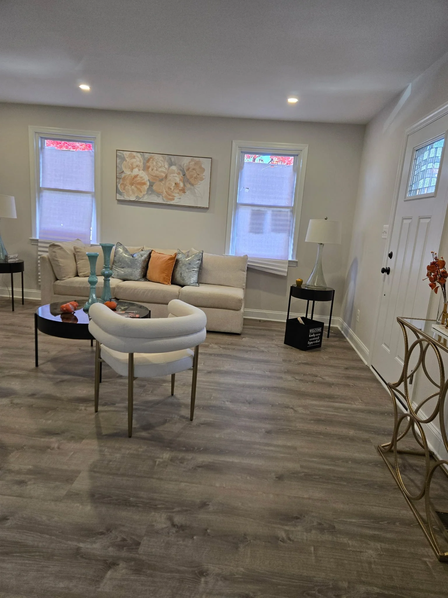 Living room with hardwood floors, a white sofa with decorative pillows, two windows with shades, a small black side table with a lamp, and a round coffee table with vases and decorative objects.