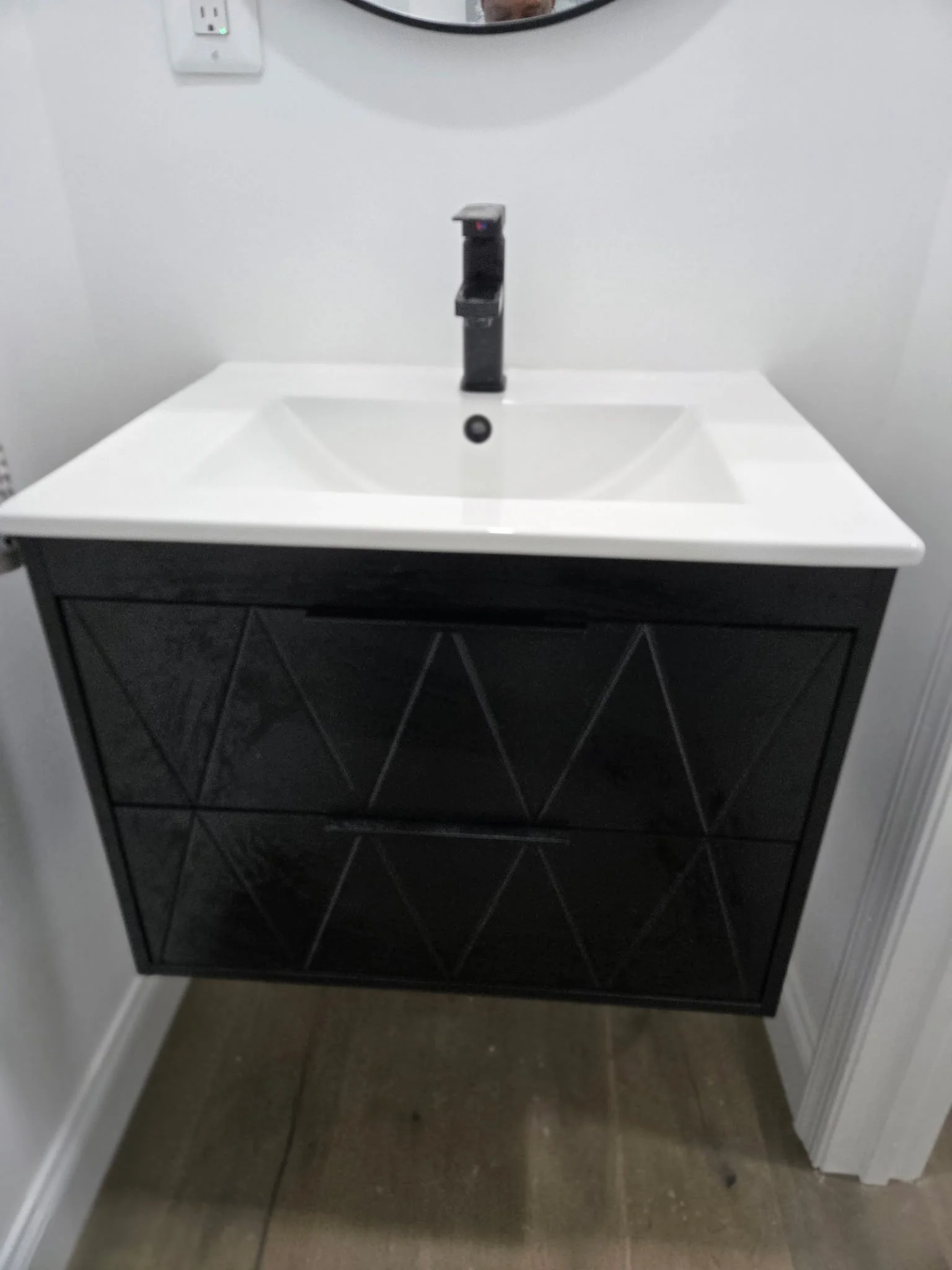A modern bathroom vanity with a white sink and black cabinet with geometric patterns, mounted on the wall, with a round mirror above and a wall outlet nearby.