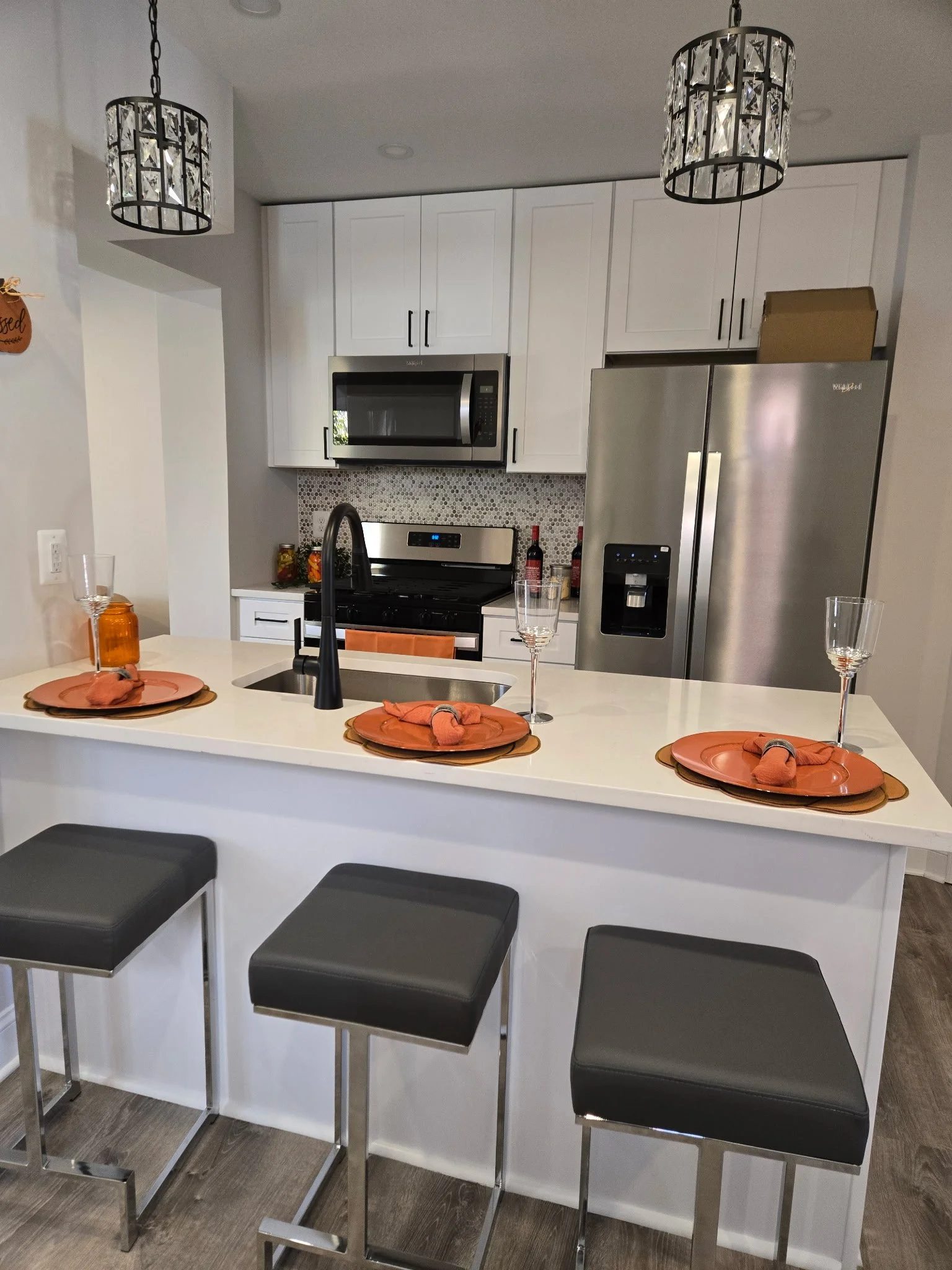 Modern kitchen with white cabinets, stainless steel appliances, black and white backsplash, and a kitchen island with three black cushioned stools. The island is set for three with orange placemats, matching napkins, and clear wine glasses.