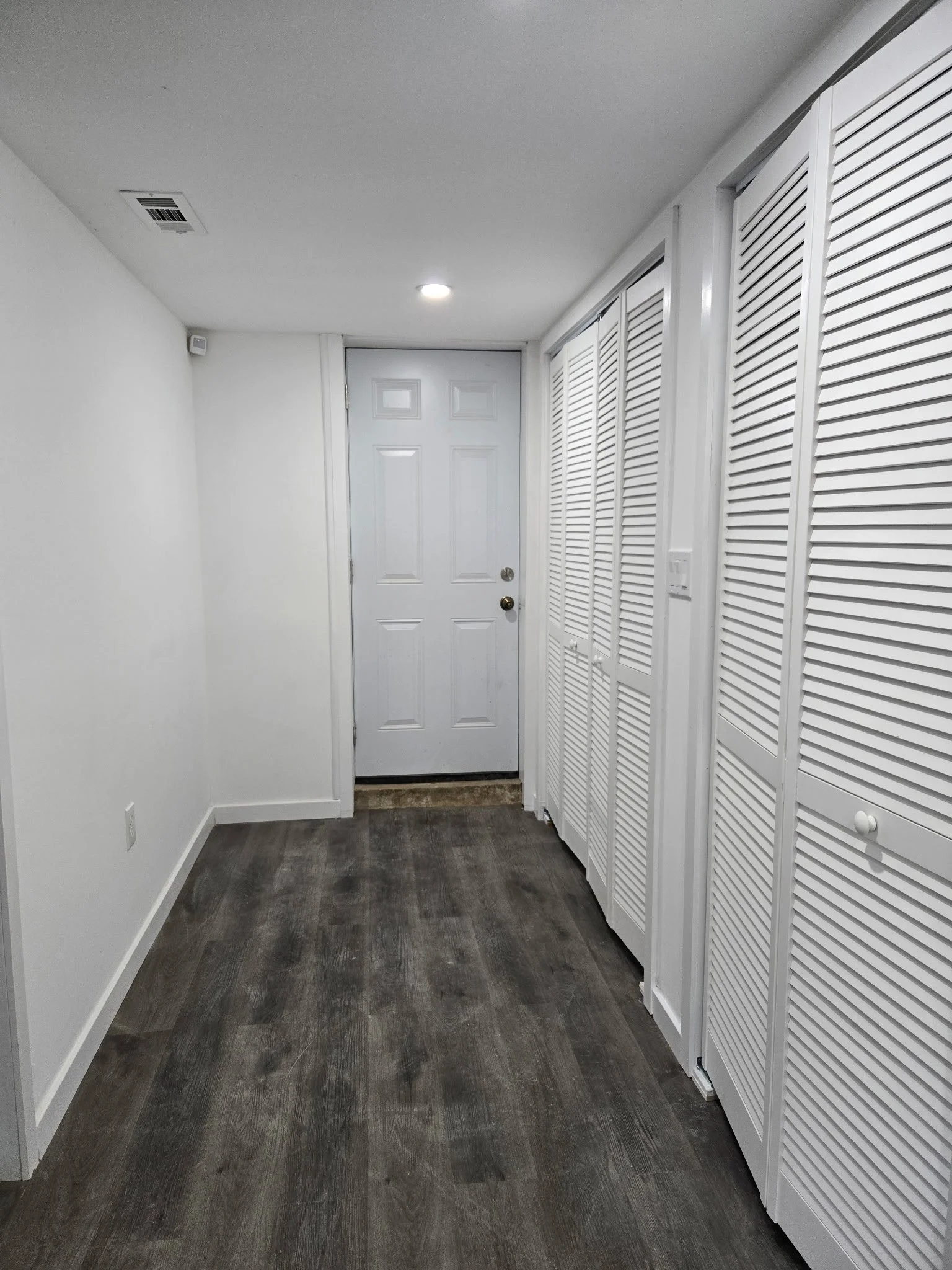 Empty hallway with white walls, wooden flooring, white louvered closet doors on the right, and a white door at the end of the hall.