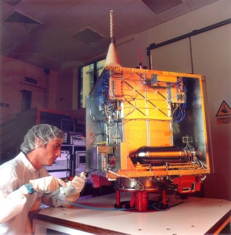 A scientist in a laboratory wearing a hairnet, gloves, and a white lab coat working on a small spacecraft or satellite.