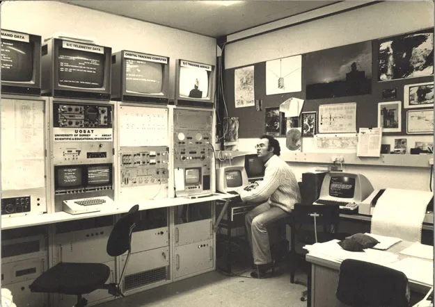 A man sitting at a desk in a control room with multiple old computer monitors, control panels, and equipment, surrounded by various pictures and information posters on the walls.