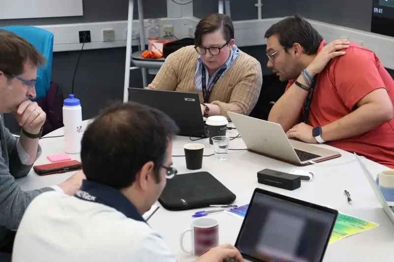 Group of five people working together at a meeting table, analyzing documents and using laptops.