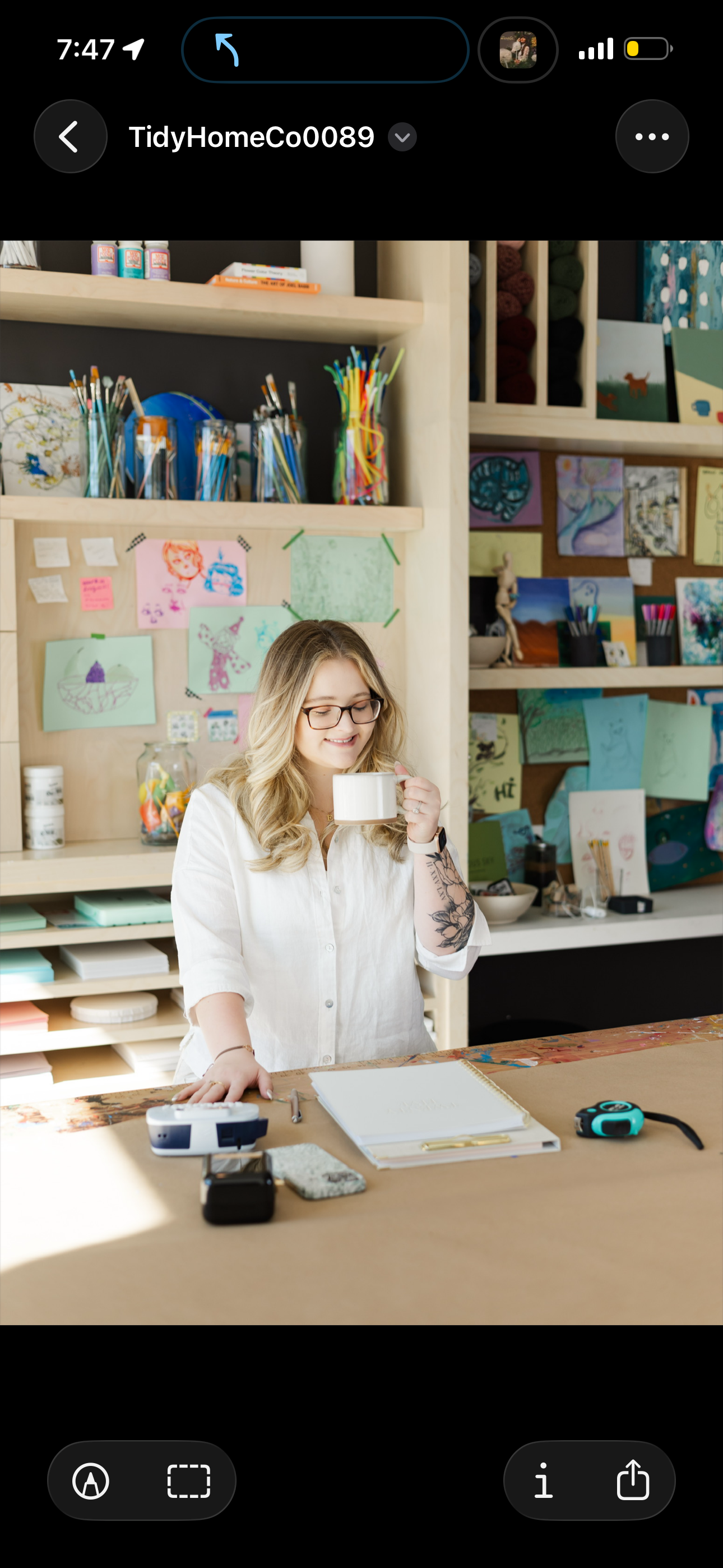 A woman with glasses and tattoos smiling while holding a white mug in a colorful art studio.