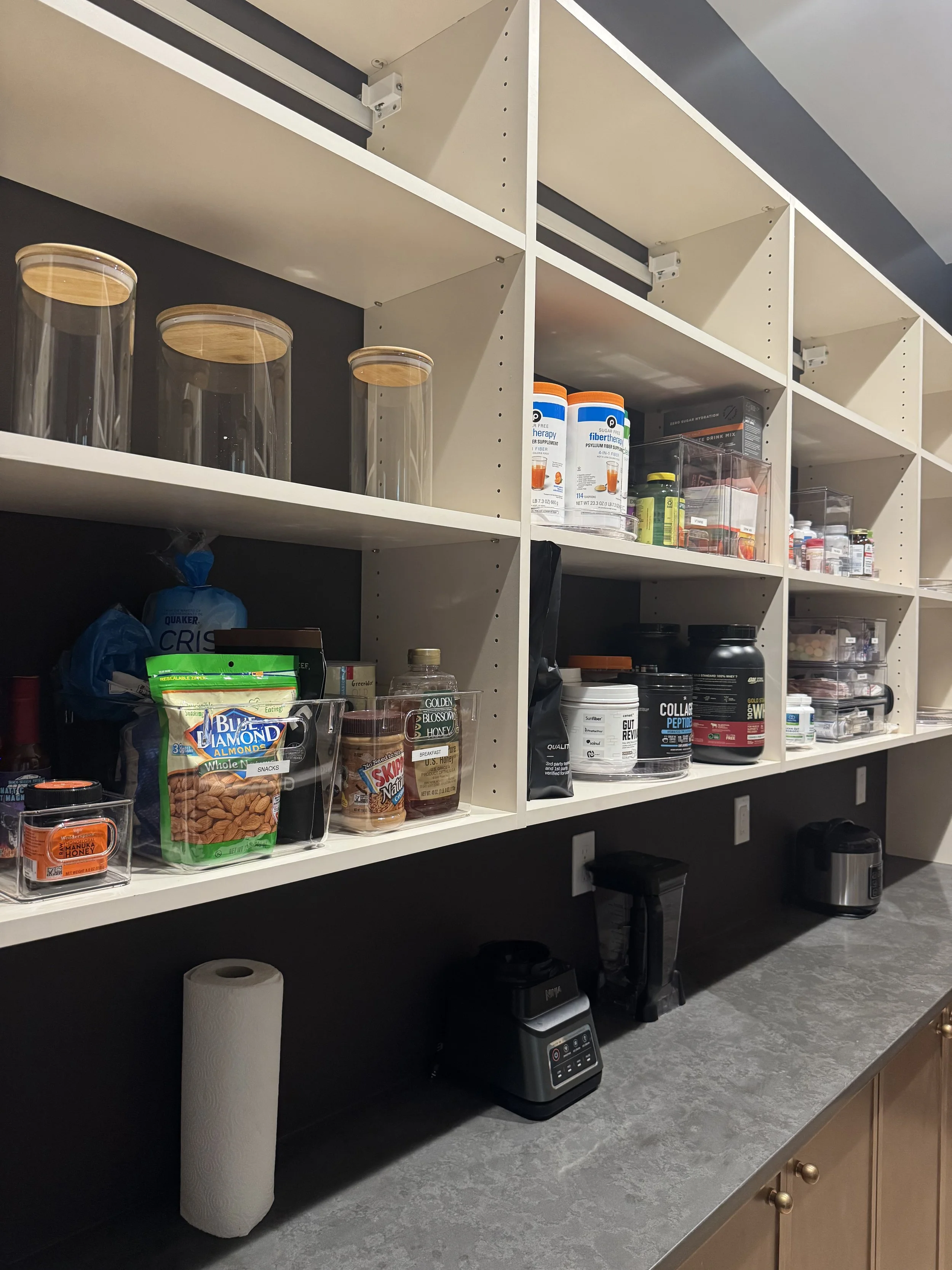 Kitchen or pantry with open white shelves containing food supplements, containers, and snacks. On the counter are a paper towel roll, a blender, and a black appliance.