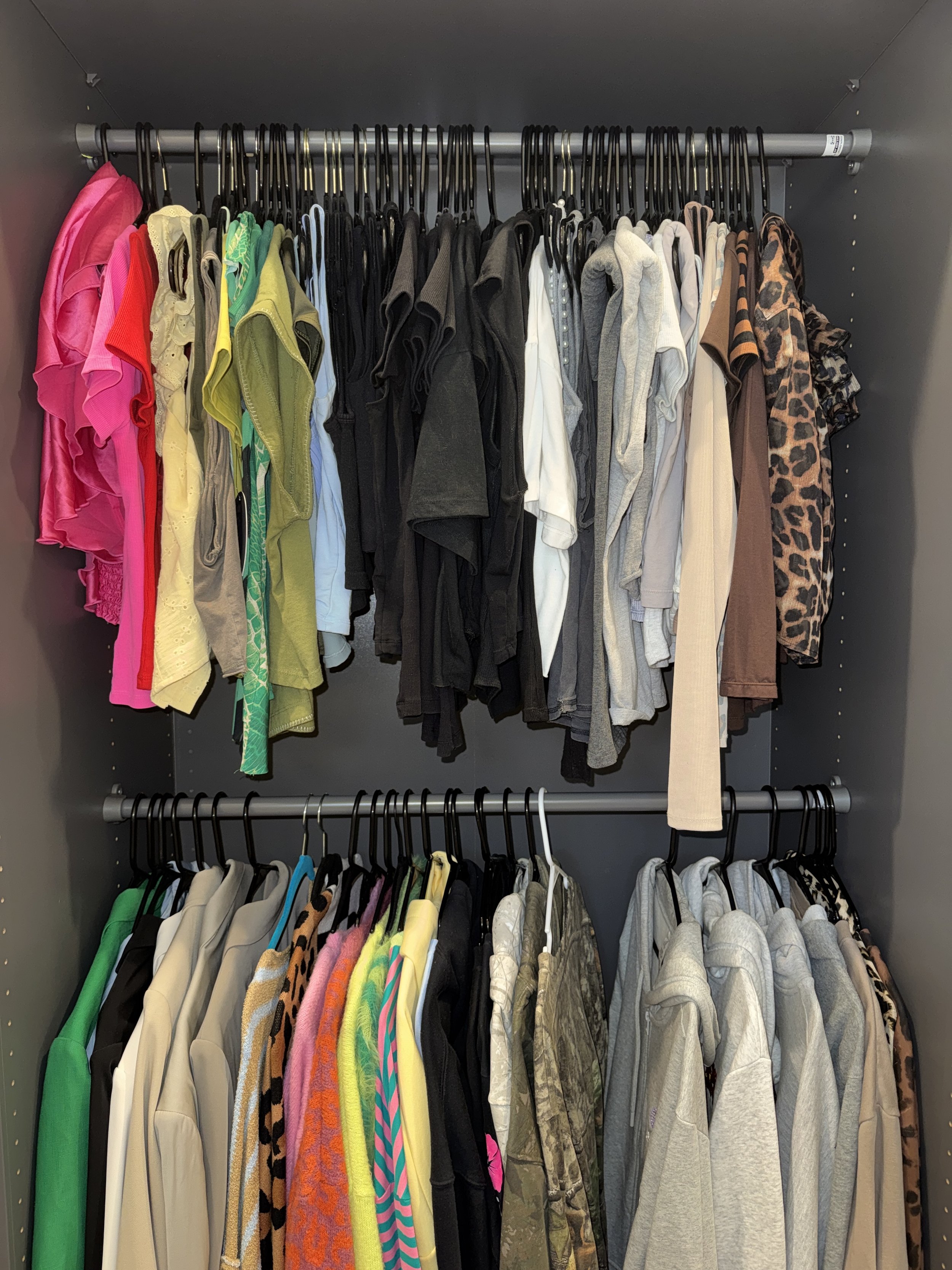 A closet filled with hanging clothes, including colorful shirts, hoodies, jackets, and pants organized on black hangers on two rods.