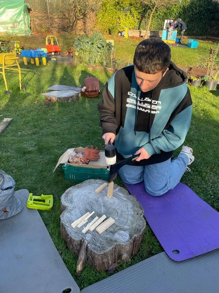 A boy kneeling on a purple mat outdoors using a handheld cordless drill to assemble wooden pieces on a large tree stump. The background features a grassy yard with children's toys, garden beds, and a person working near a blue container.