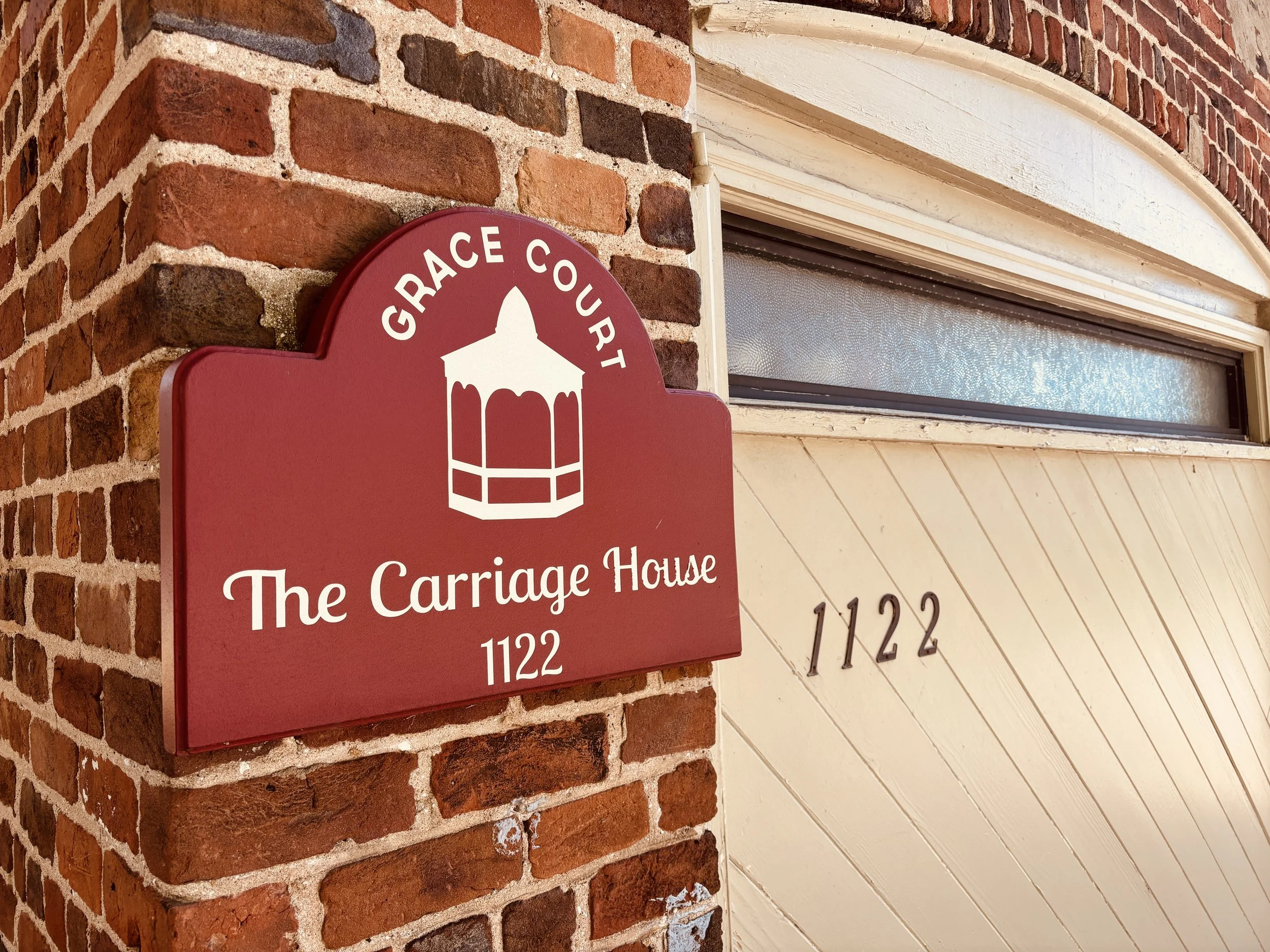 A red sign with white text and a gazebo icon reading: "GRACE COURT The Carriage House" and the number 1122 on the brick wall above a window.