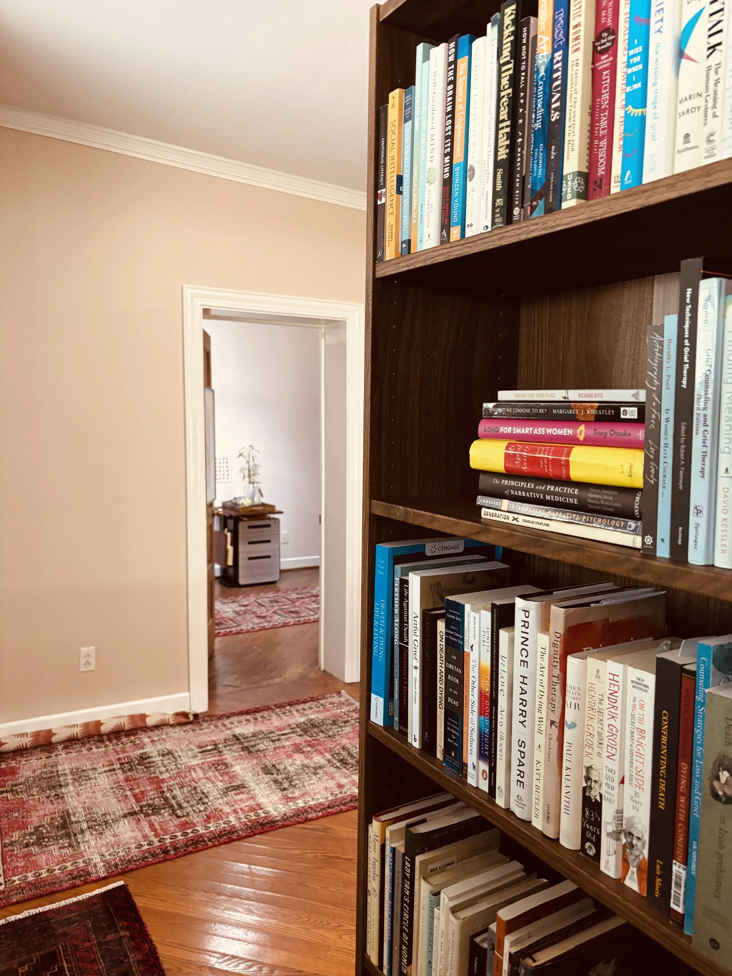 A view of a room with a wooden bookshelf filled with books and a doorway leading to another room with a desk and plant visible. The room has hardwood floors and a patterned red rug.