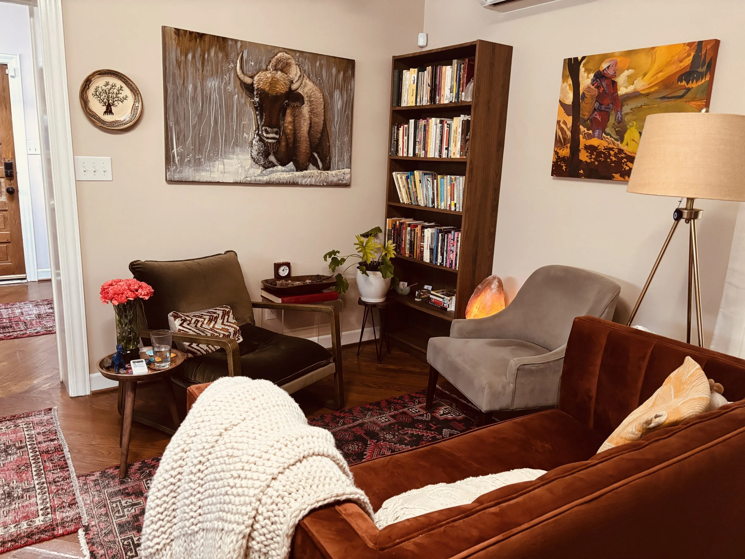 Living room with paintings of a bison and a woman on a mountain trail, bookshelf, armchairs, a salt lamp, and various decorations.