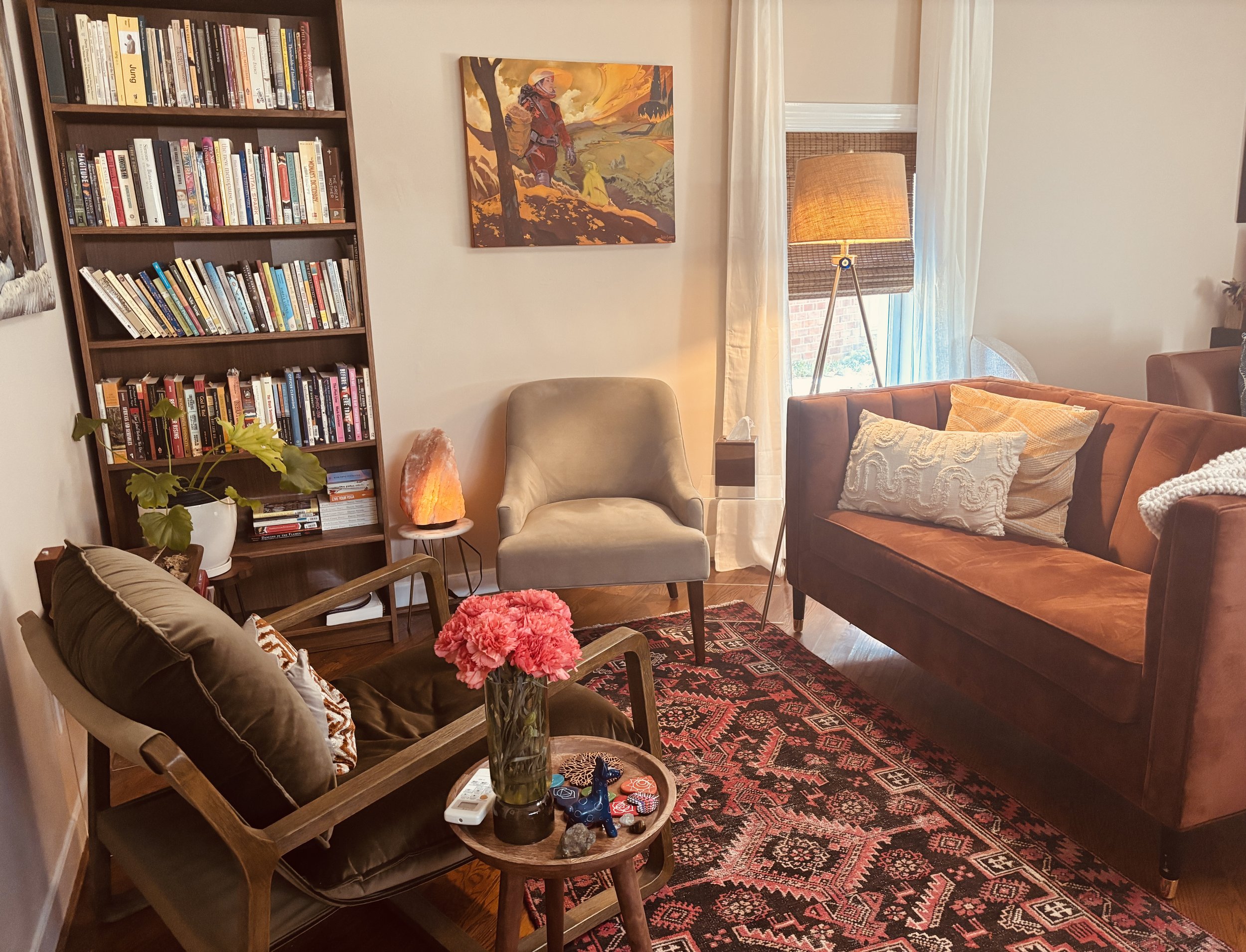 Living room with a bookshelf filled with books on the left, a beige chair and salt lamp next to it, a colorful painting on the wall, a window with white curtains, a tripod lamp, a brown couch with decorative pillows, a red patterned rug, and a small table with flowers and decorative items.