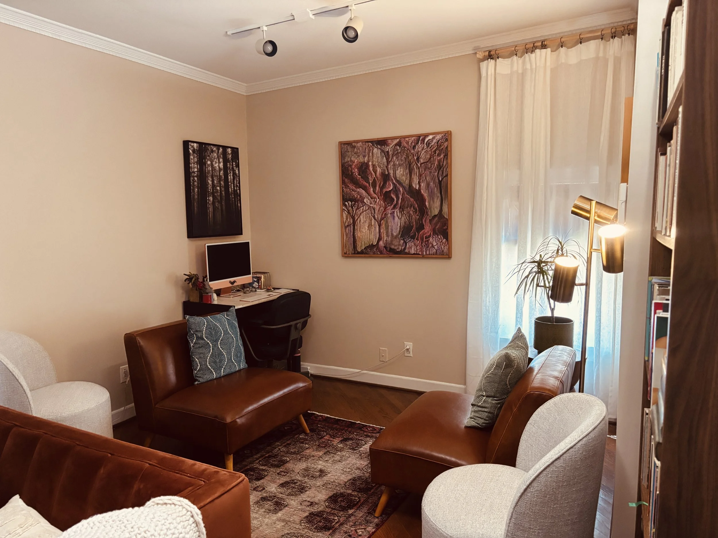 Living room with a beige wall, a painting of a forest on the wall, a desk with an iMac computer, seating including brown leather and white upholstered chairs, a window with sheer curtains, a floor lamp, and a wooden bookshelf.
