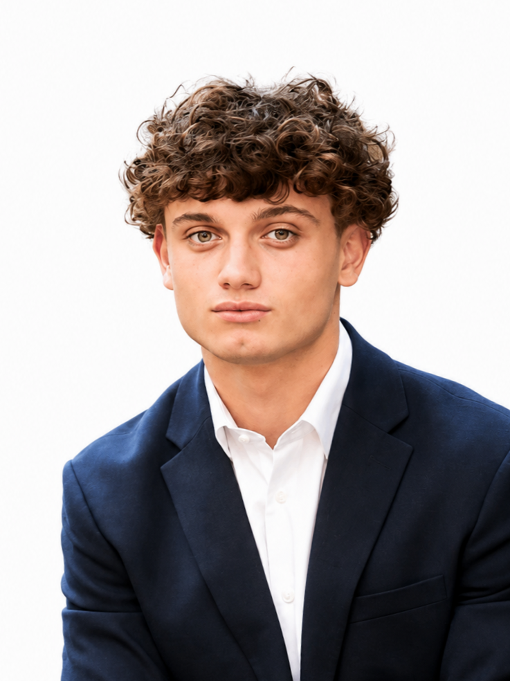 A young man in a navy blue suit and white shirt sitting outdoors with green leaves in the background.