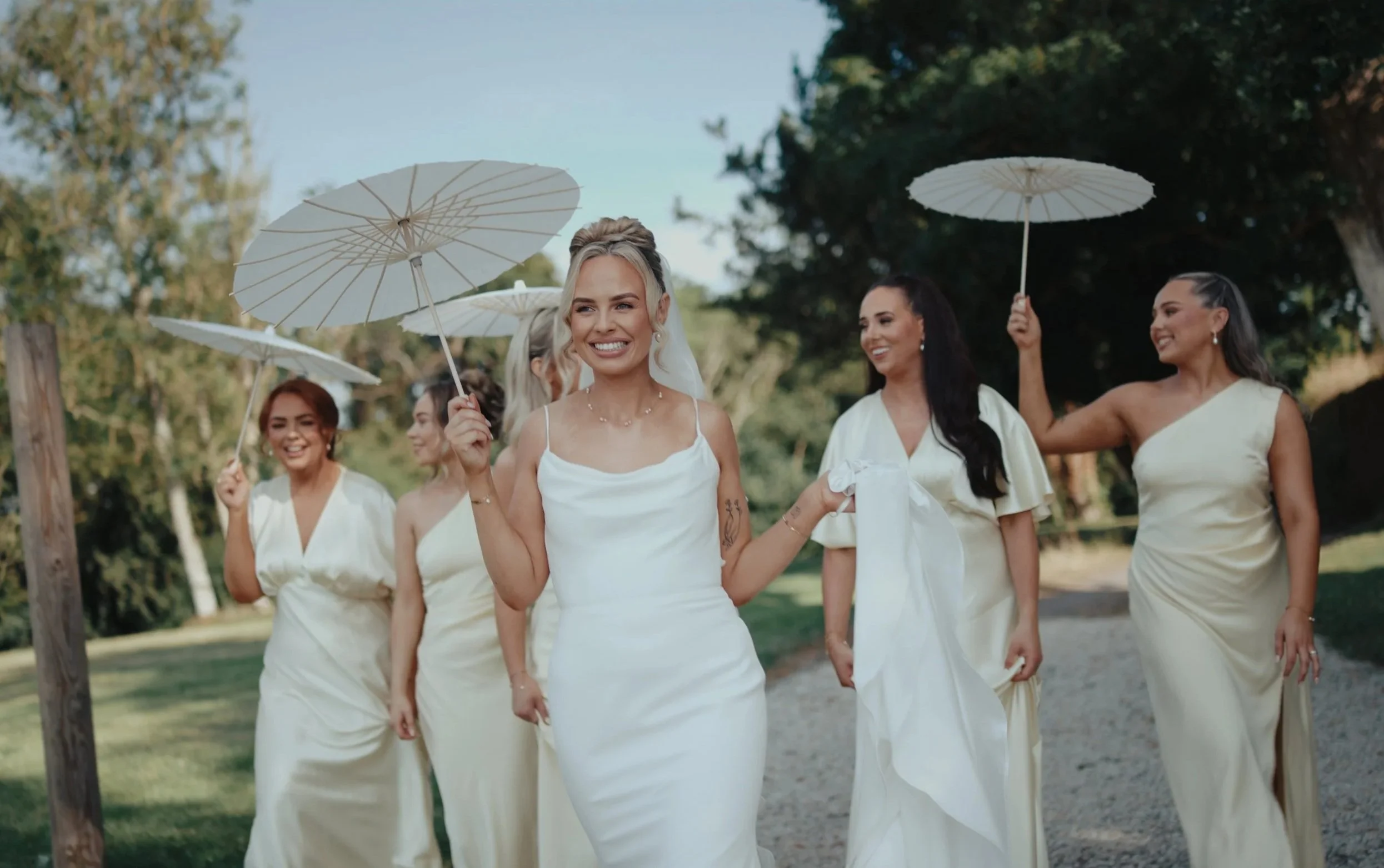 A group of women in white dresses holding white parasols walking outdoors in a natural setting.