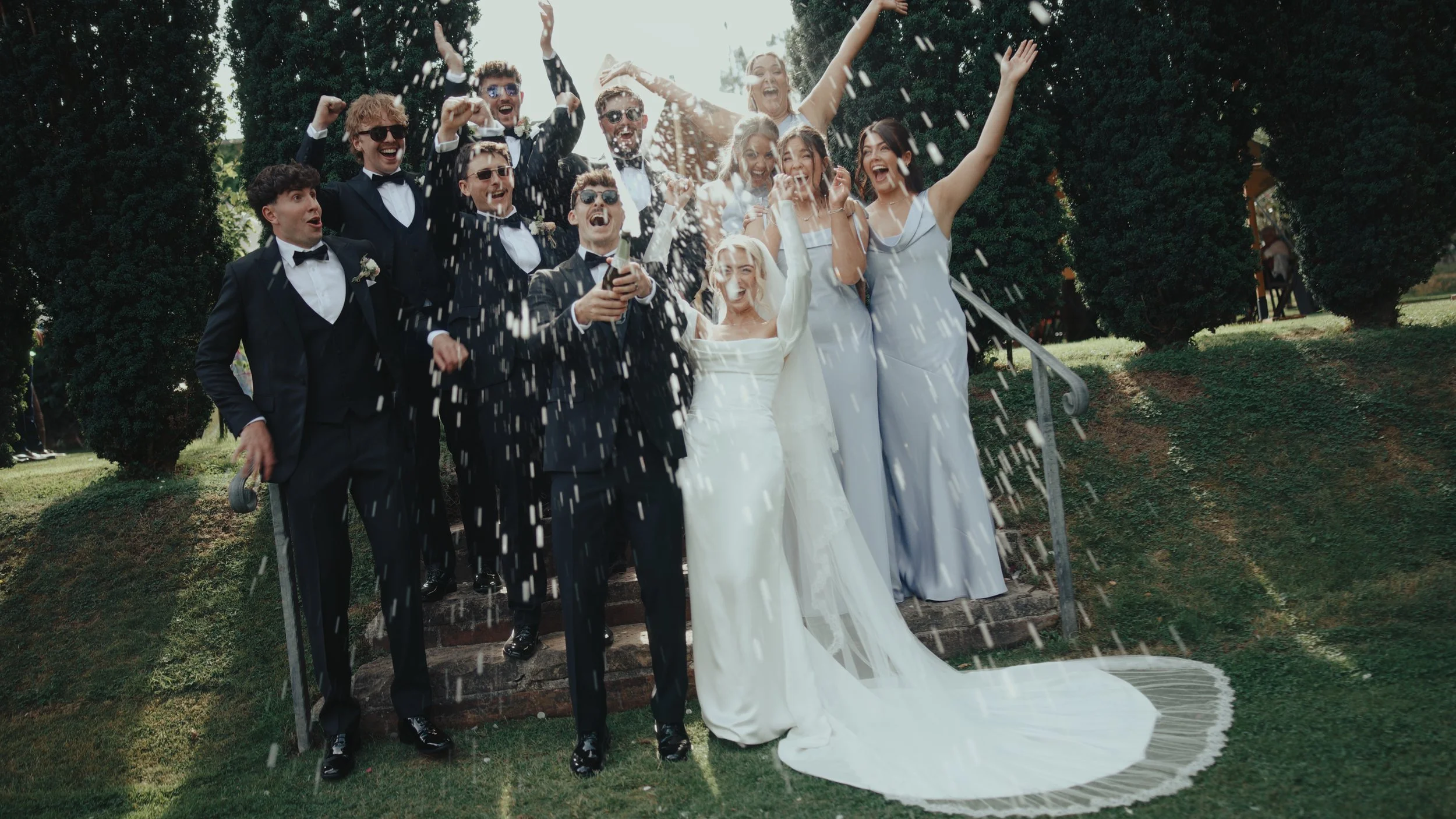 Group of newly married couple and friends celebrating outdoors, throwing rice or confetti, on wedding stairs.