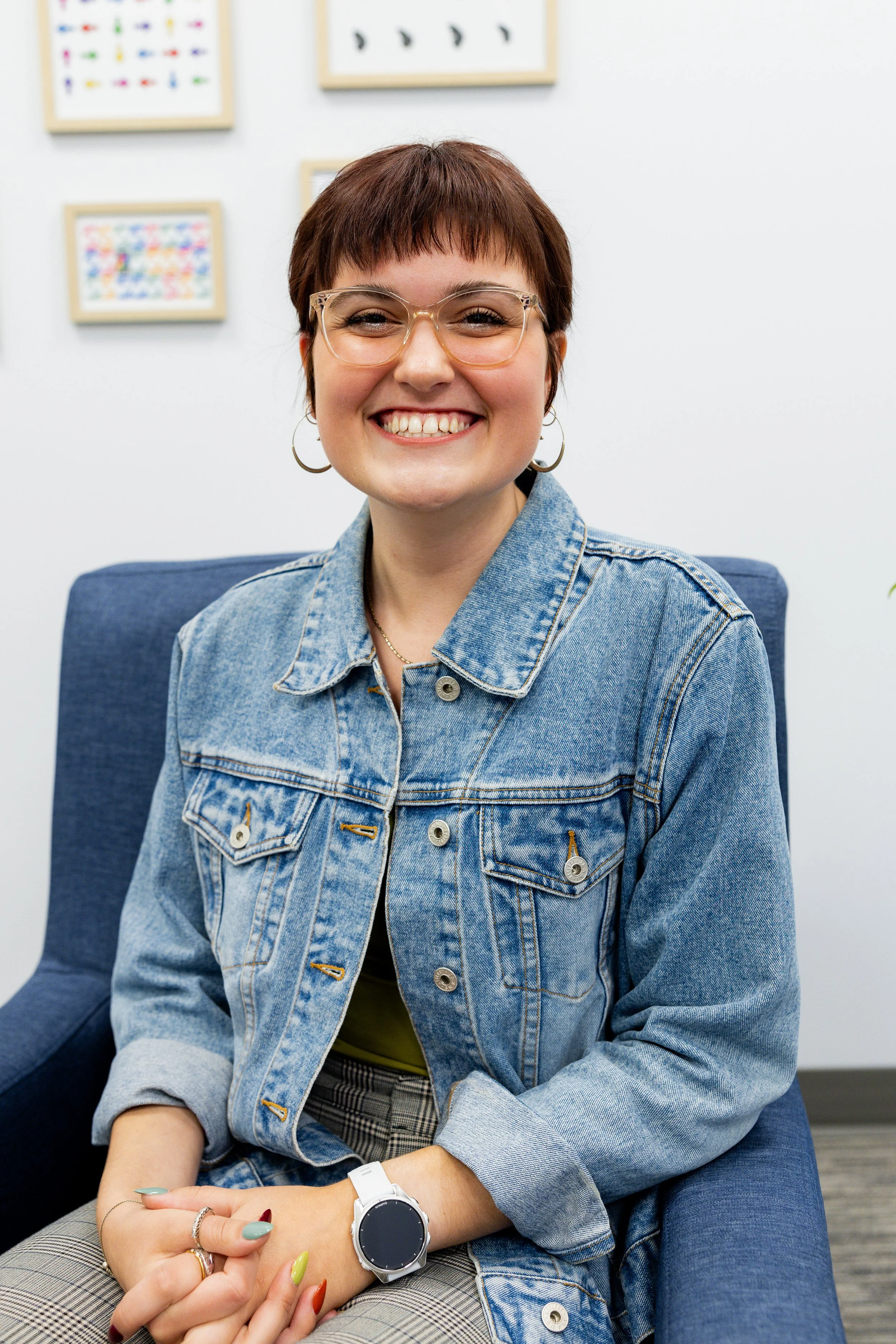 Elise, a smiling woman with short, curly brown hair and glasses seated in a blue chair in front of a wall decorated with colorful framed pictures.
