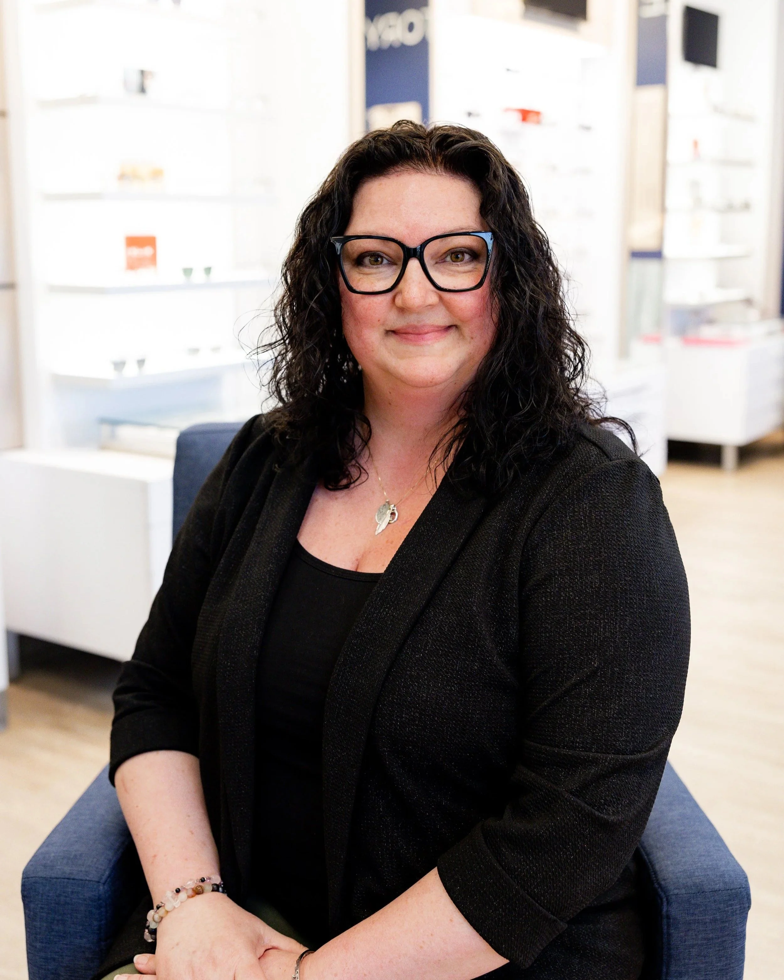 Michelle, a woman with brown hair and glasses sitting in an eye care or optical shop, smiling at the camera.