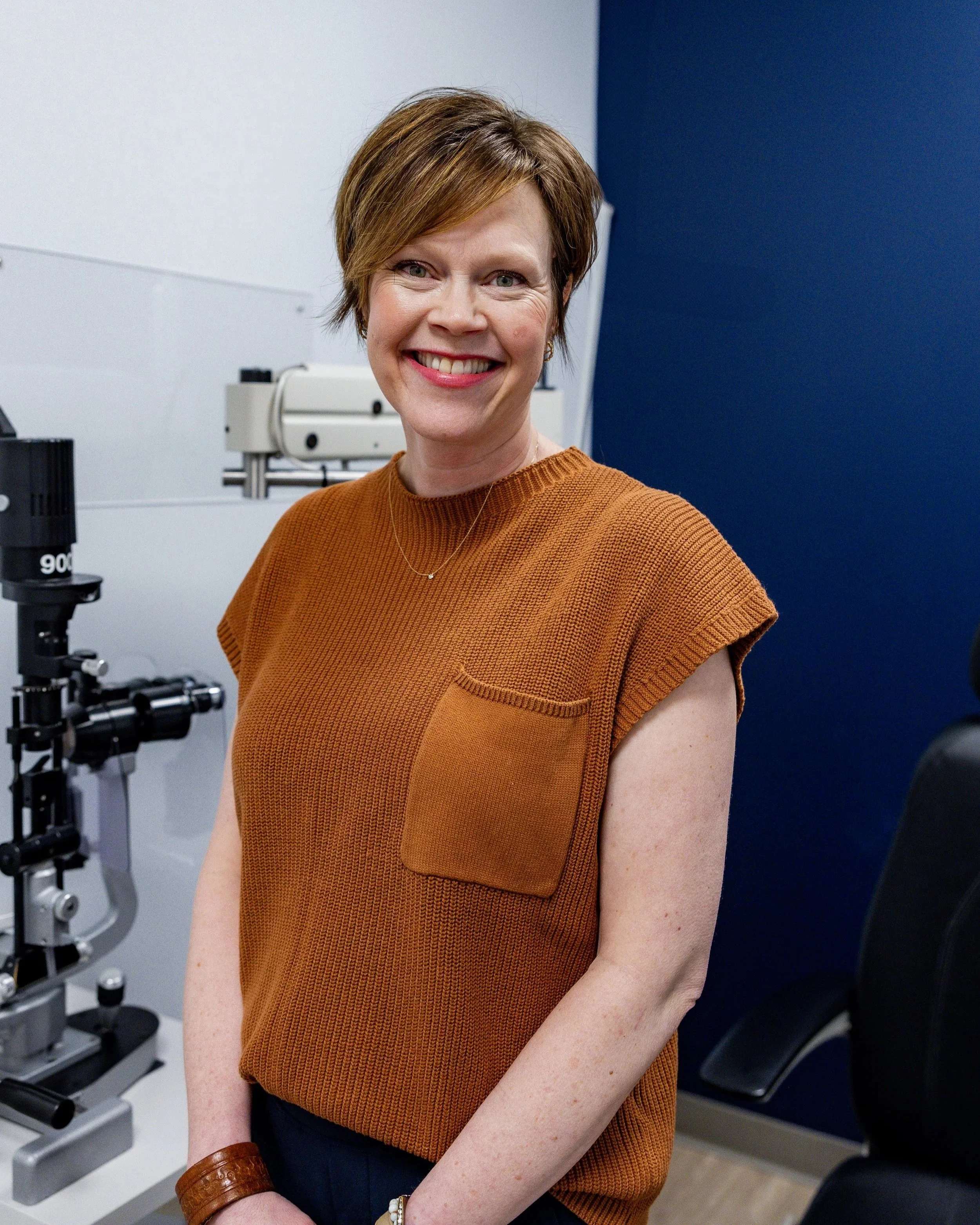 Dr. Joy Quitberg, a woman with short brown hair, wearing a white sweater and olive-green pants, is sitting on a blue chair in a medical or clinical room, smiling at the camera. In the background, there is medical equipment and a chair.
