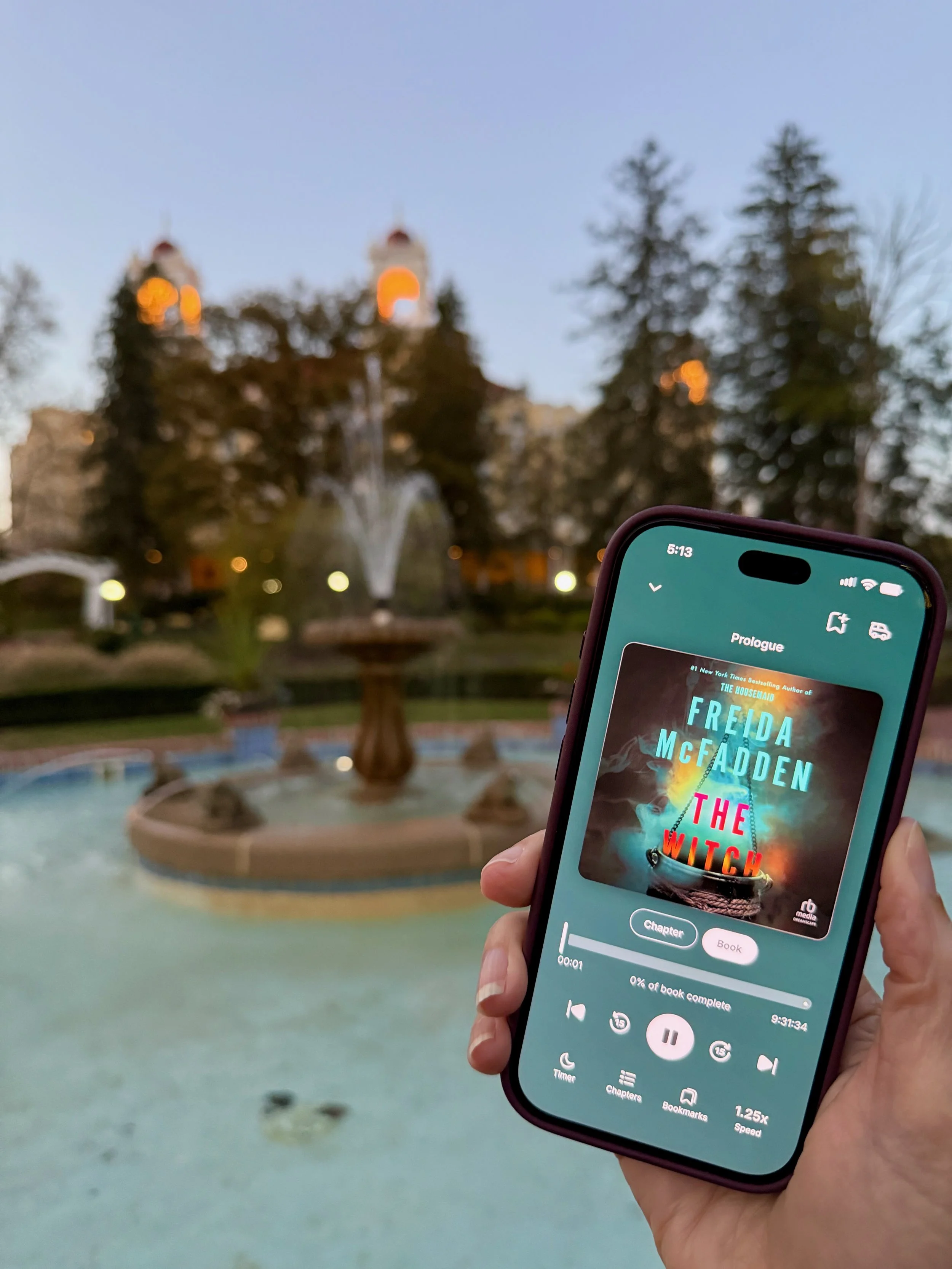 Phone displaying the audiobook The Witch by Freida McFadden, held in front of a fountain at dusk.
