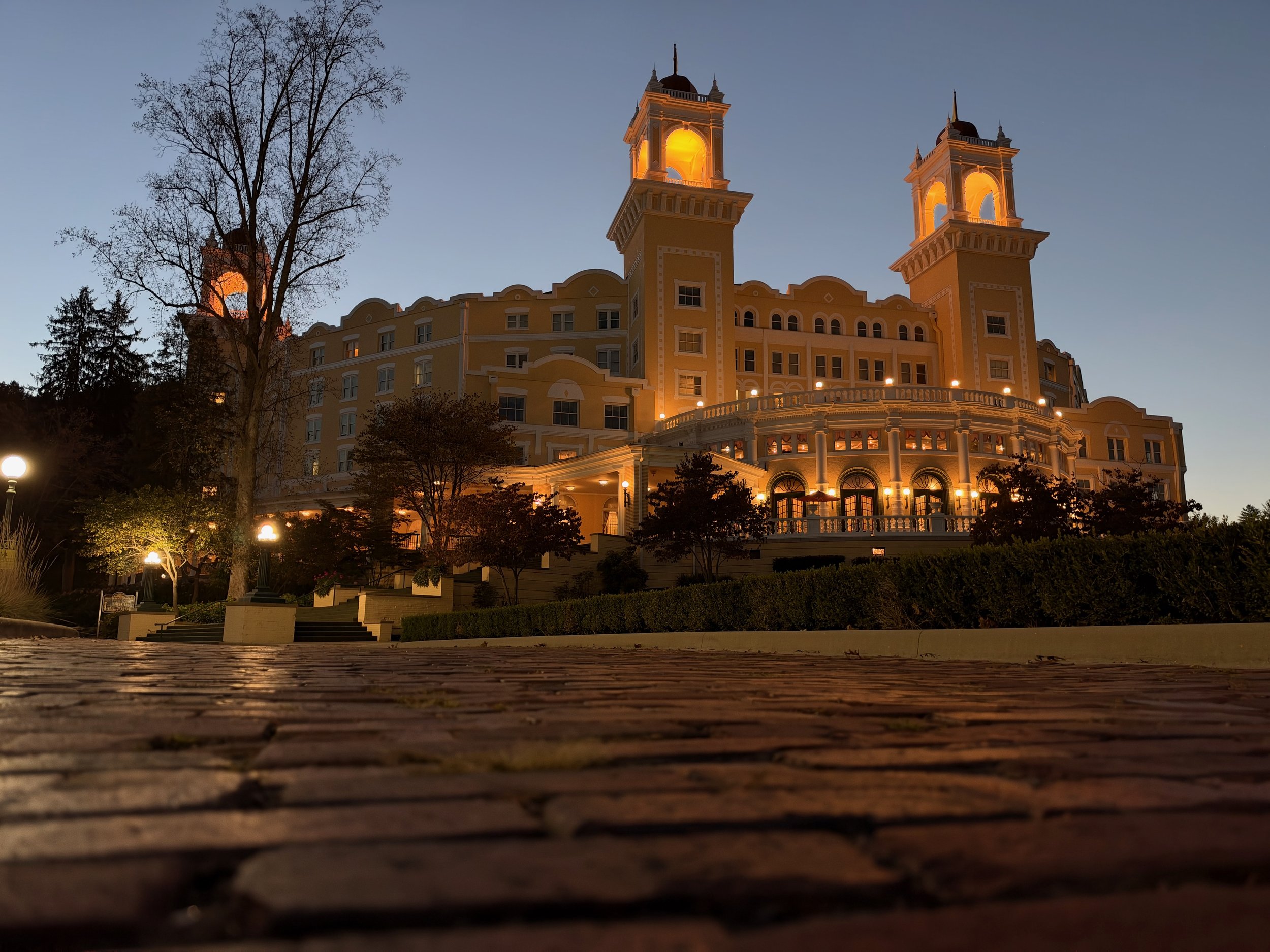 West Baden Springs Hotel glowing amber at dusk, with lit towers and a curved terrace, seen from a low angle over a red brick road.