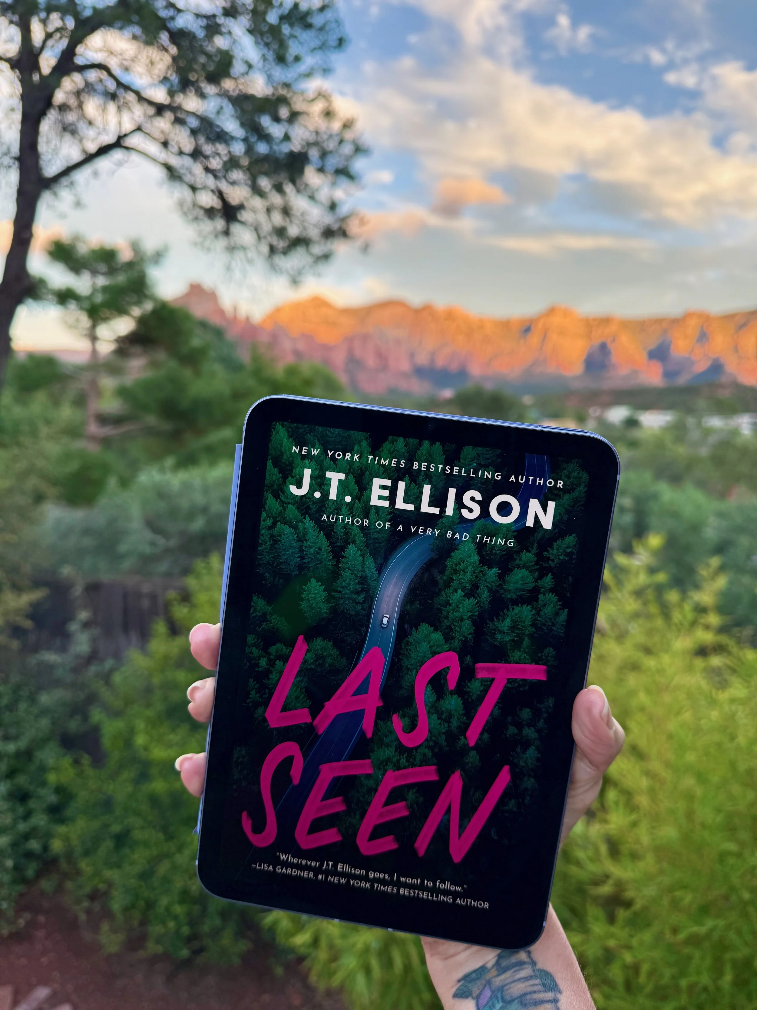 Hand holding a tablet displaying the cover of Last Seen by J.T. Ellison, with Sedona’s red rock mountains glowing in the sunset behind it and green trees in the foreground.