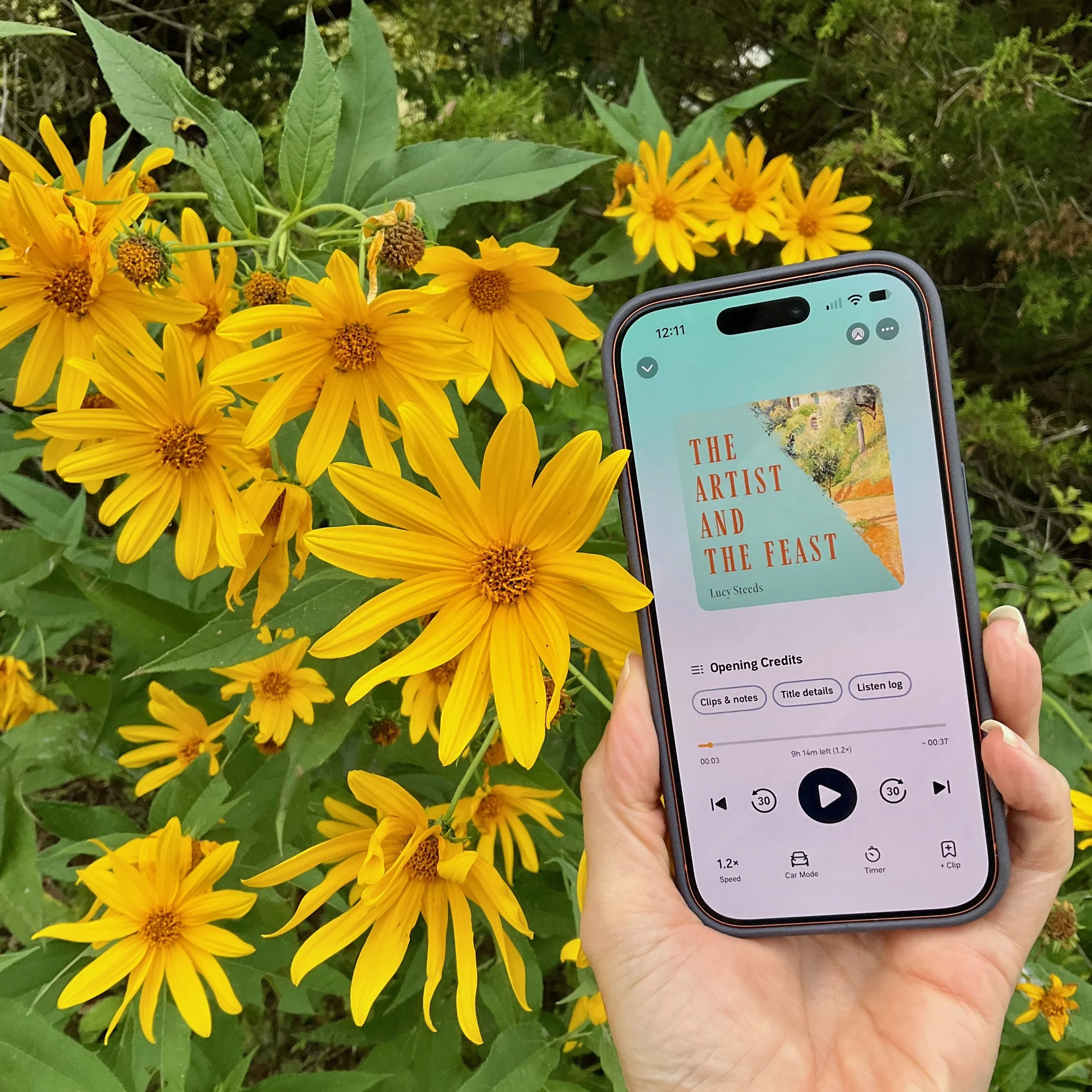 A hand holds a phone showing the audiobook The Artist and the Feast by Lucy Steeds in front of bright yellow flowers.