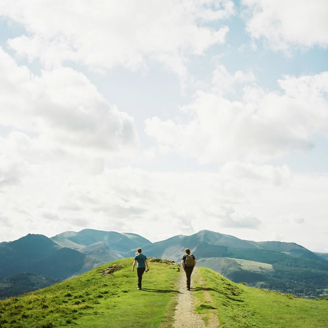 Two hikers walking on a grassy trail on a hilltop with mountains in the background under a partly cloudy sky.
