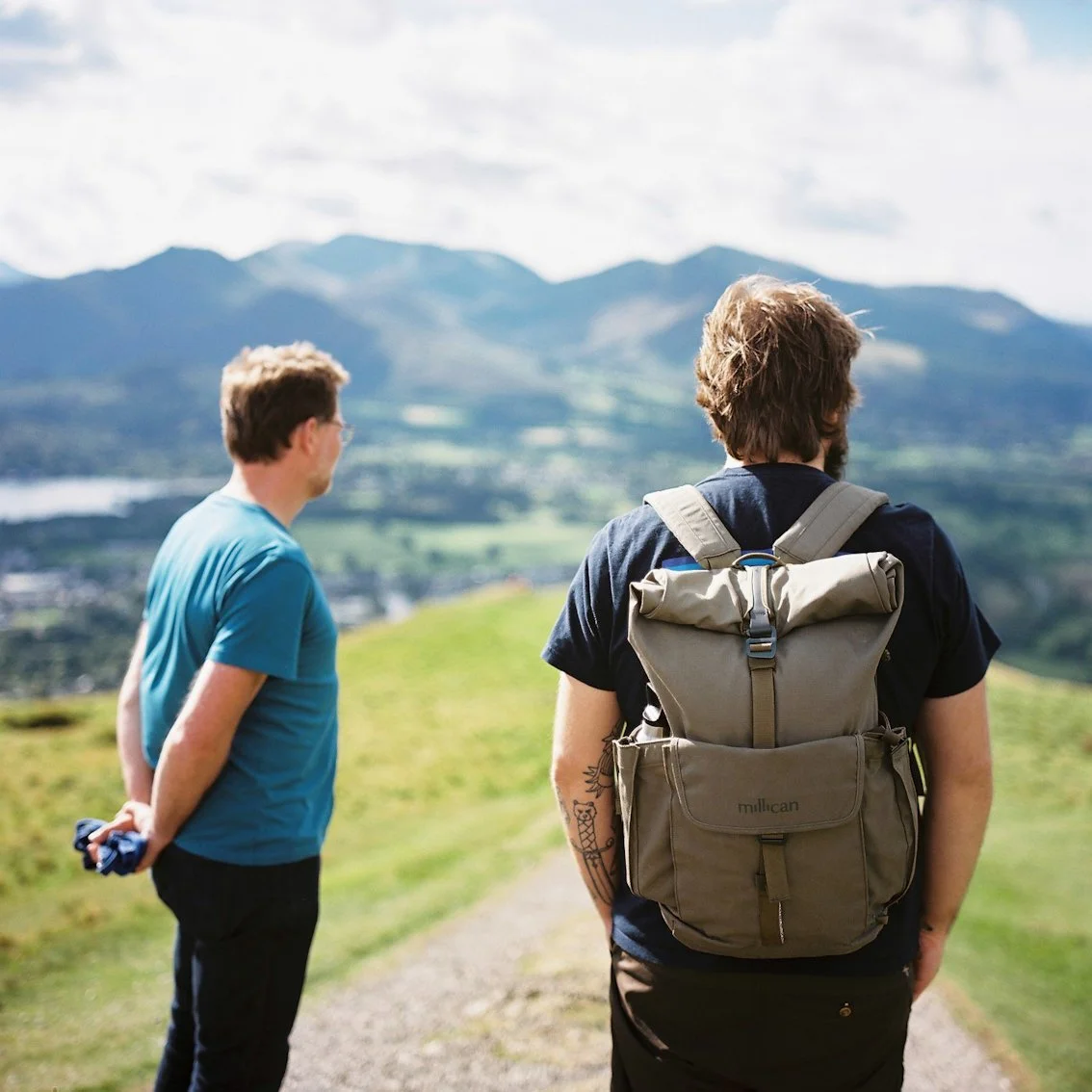 Two men standing on a grassy hill overlooking mountains and a lake, one with a backpack and the other holding a jacket behind their back, with cloudy sky above.
