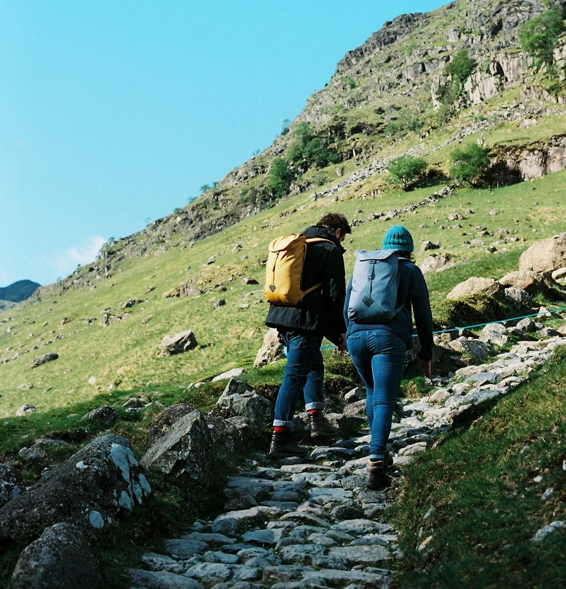 Two people with backpacks hiking up a rocky trail on a mountain slope with greenery and a clear blue sky.