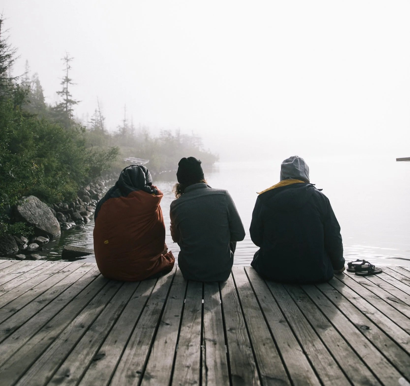 Three people sit on a wooden dock by a foggy lake, with trees in the background, facing the water.