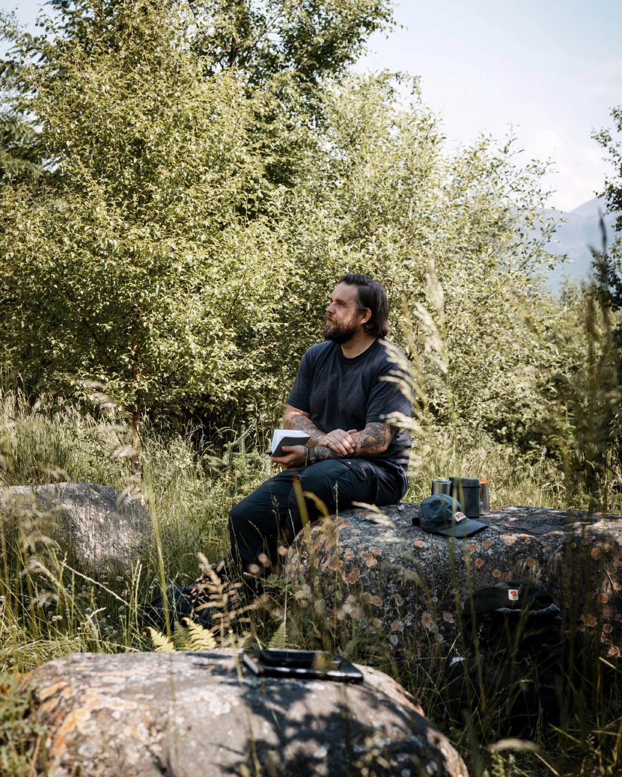 A man with dark hair and tattoos sits on a log in a lush green forest, holding a notebook, with camping gear nearby.