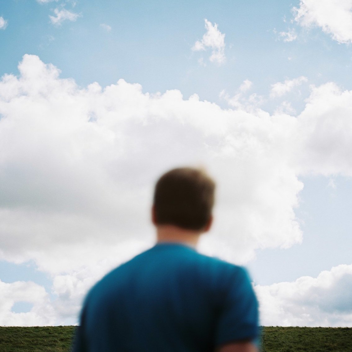A person with short hair wearing a blue shirt standing outdoors and looking up at a sky filled with fluffy clouds.