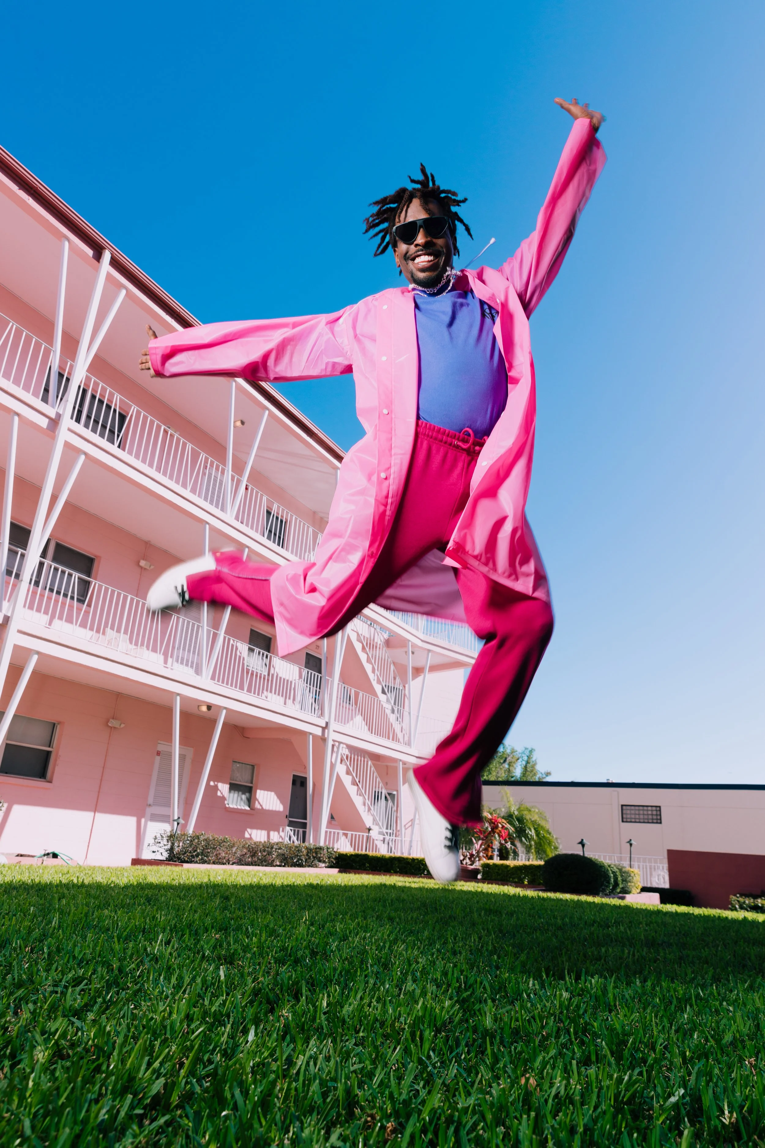 A person jumping in the air with arms outstretched, wearing a pink jacket, pink pants, a purple shirt, white sneakers, and sunglasses, outdoors near a pink apartment building under a clear blue sky.
