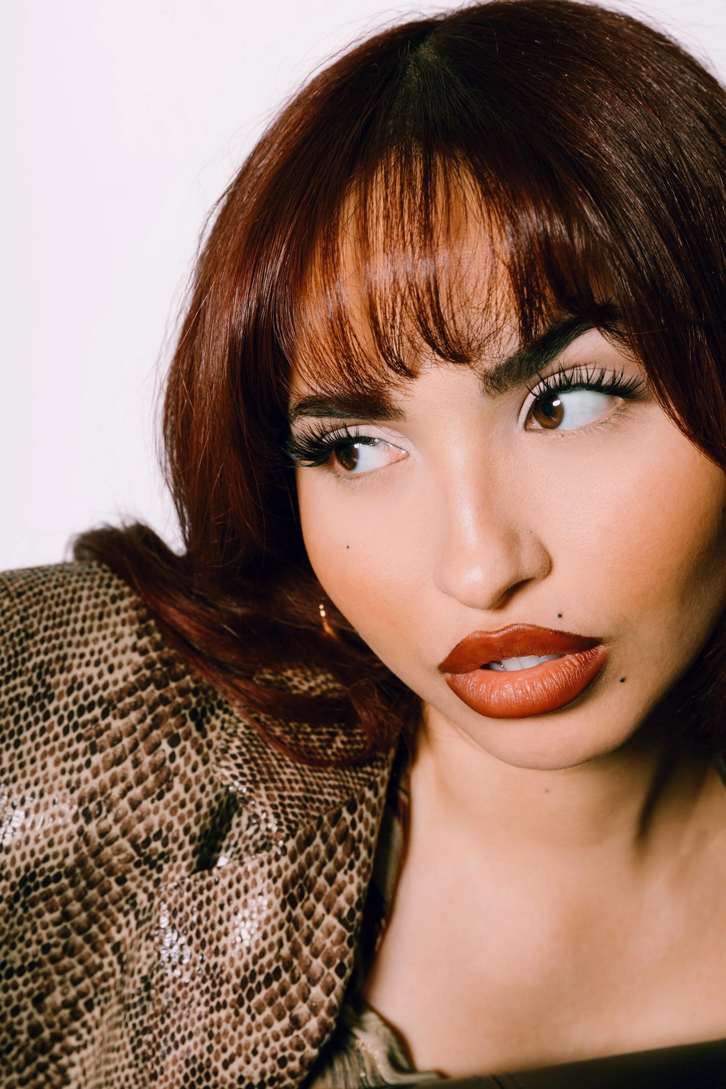 Close-up of a woman with auburn hair, brown eyes, bold makeup, and wearing a snakeskin patterned jacket, looking to the side.