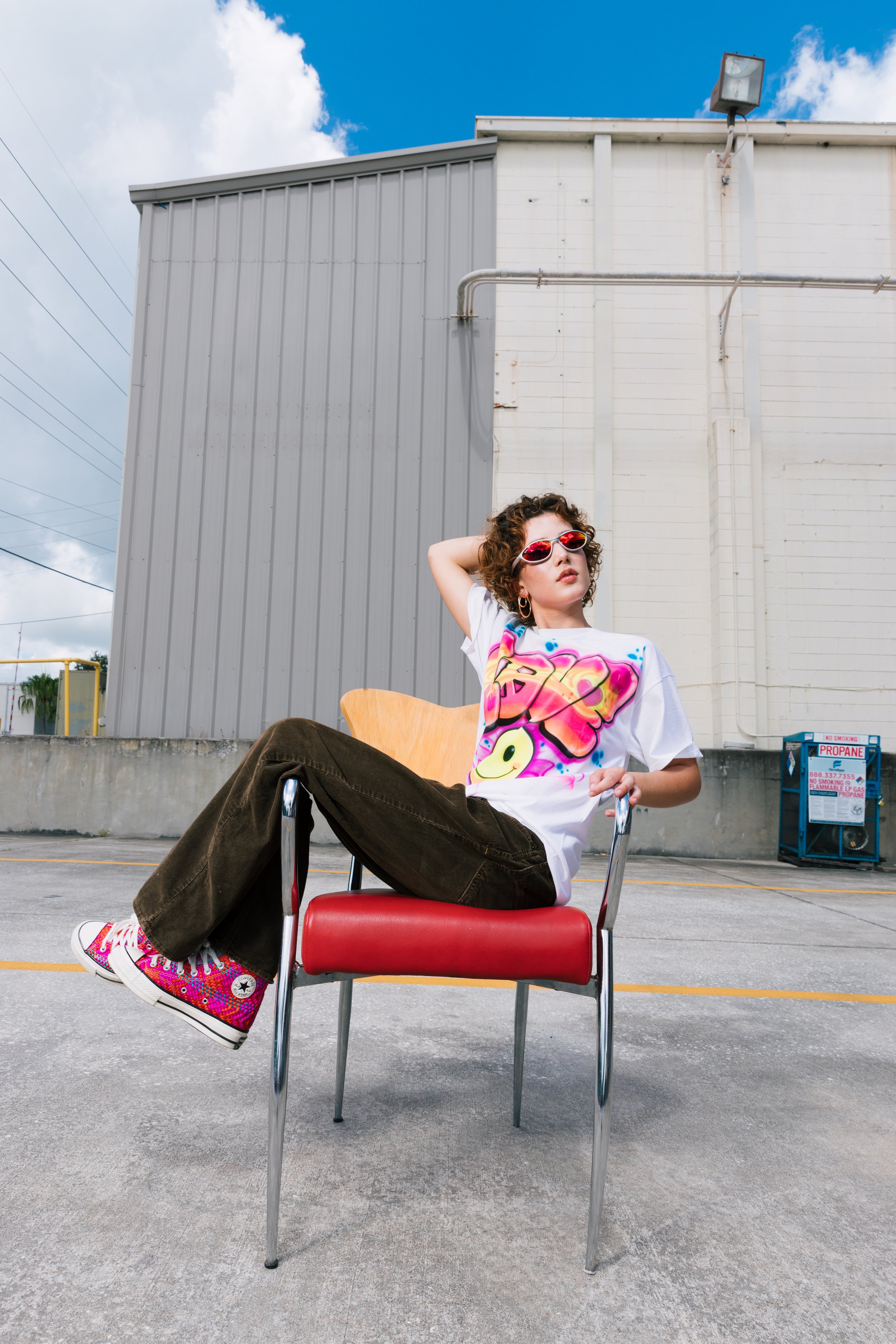 A young woman with curly hair and pink sunglasses sitting in a red chair outdoors against a background of a gray and white building, with her legs crossed on the chair, wearing a white graphic T-shirt, dark pants, and colorful sneakers.