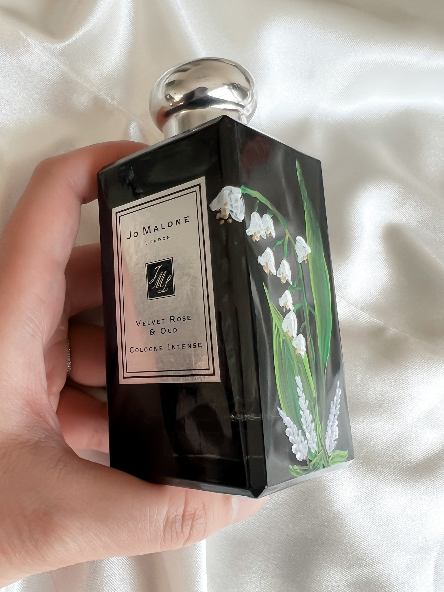 A person holding a black perfume bottle with a silver rounded cap, featuring a lily of the valley flower design and a label that reads 'Jo Malone London Velvet Rose & Oud Cologne Intense.' The background is a shiny, cream-colored fabric.